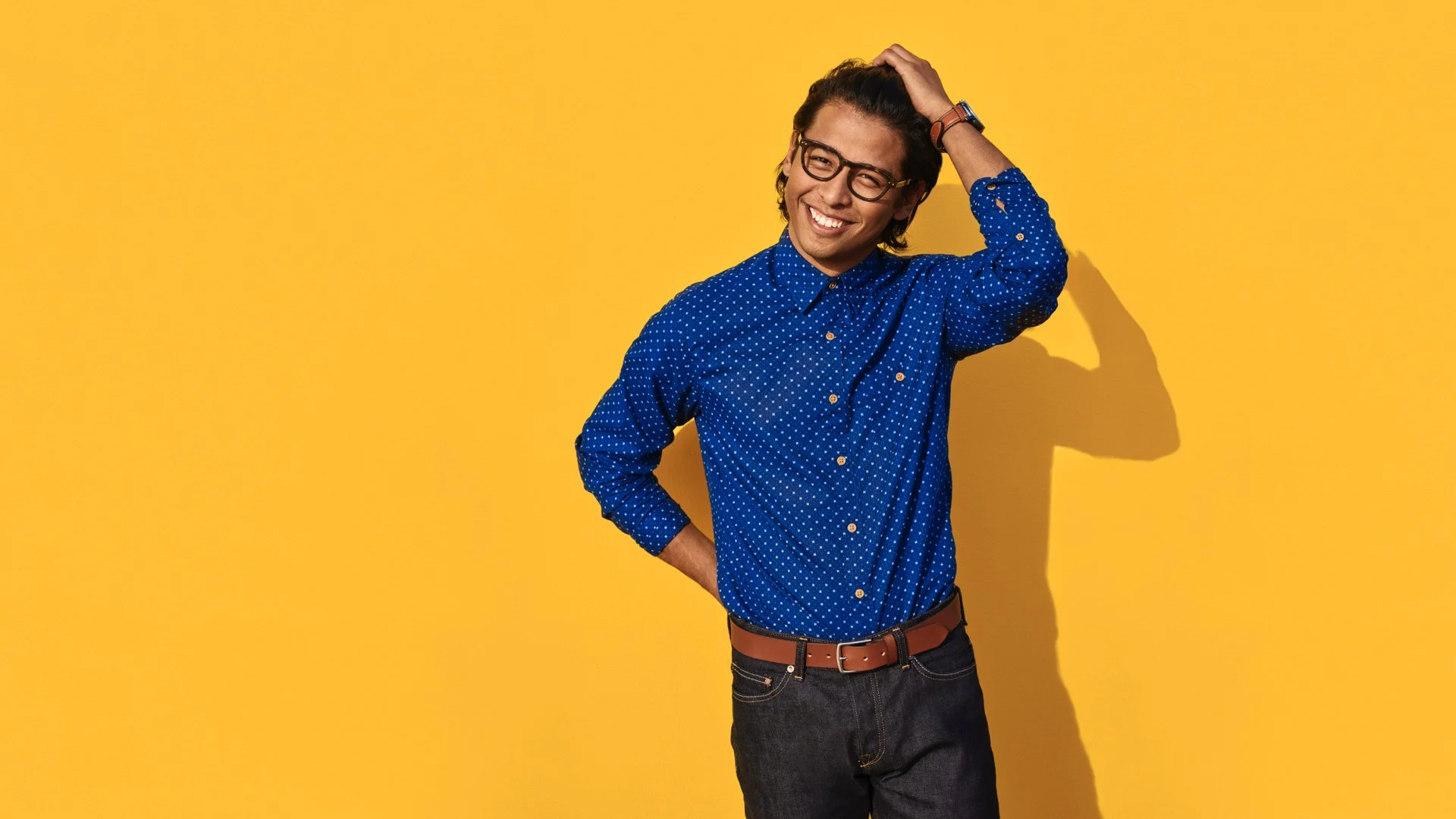 A young man with glasses, wearing a blue polka dot shirt, posing smiling with one hand on his head against a yellow background.