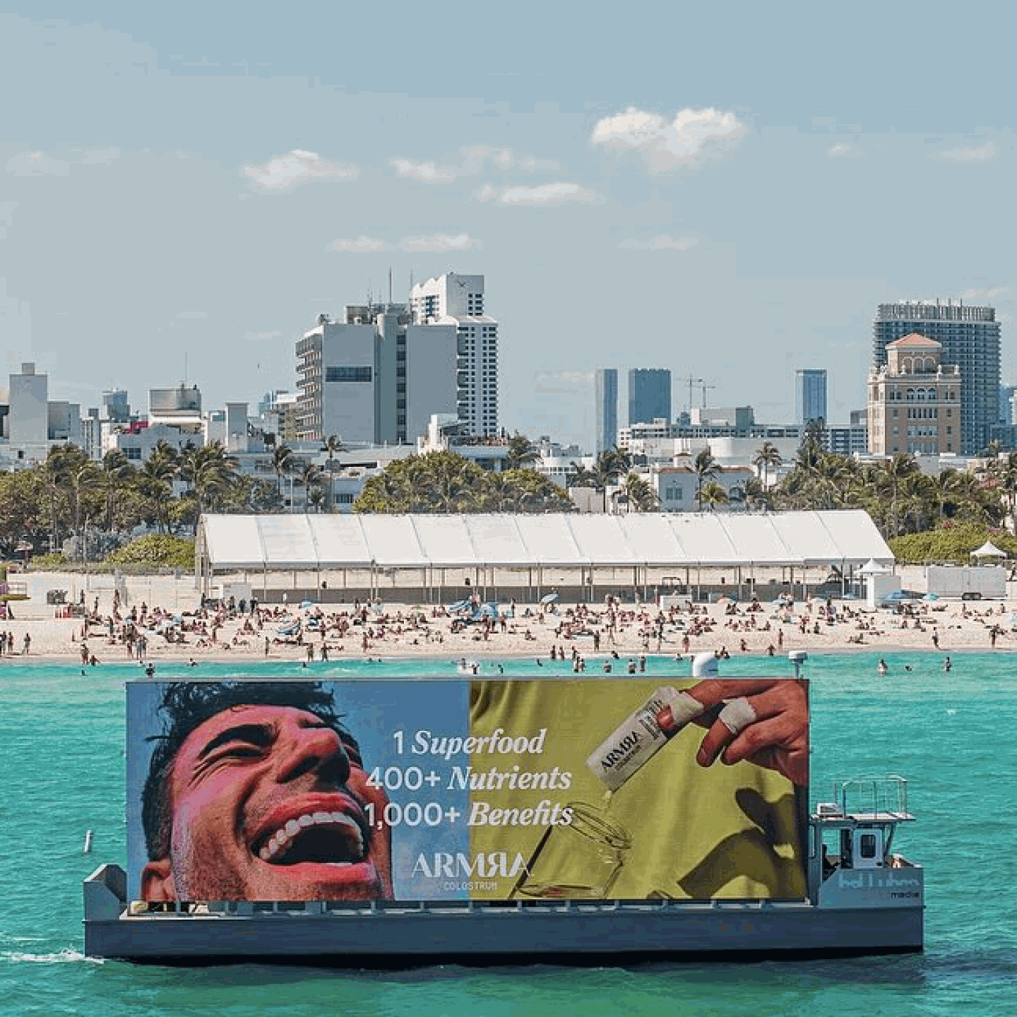 A boat with an advertisement on it floating in turquoise water near a beach with many people, with city buildings and palm trees in the background.