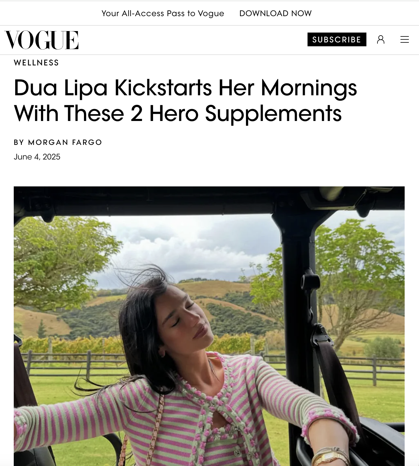 Dua Lipa with eyes closed, wearing a pink and green striped sweater, sitting in a golf cart outdoors with green trees and a hilly landscape in the background.