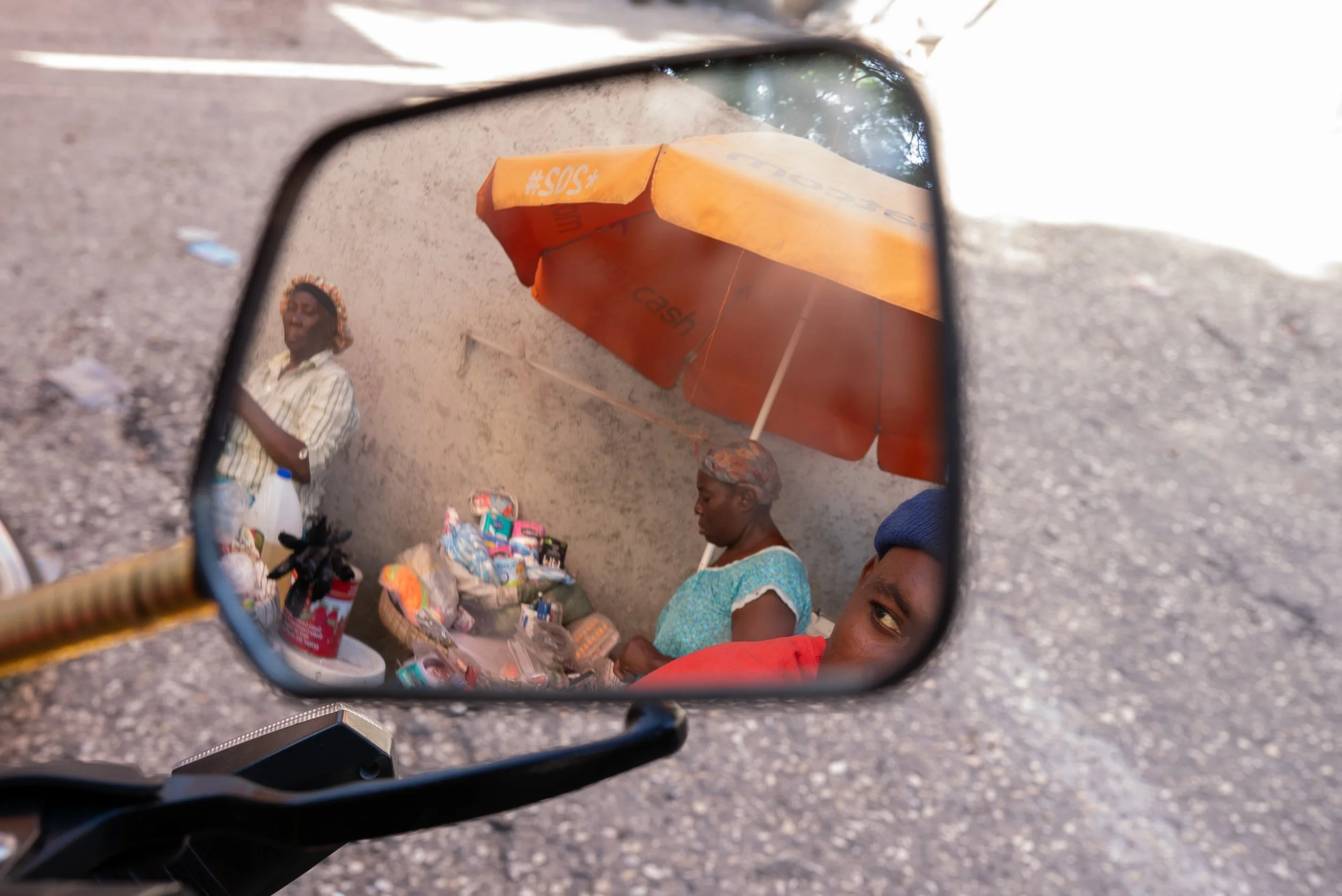 A motorcycle driver watches passerby and marketwomen as he waits for a client in the streets of Port-au-Prince. 2022.