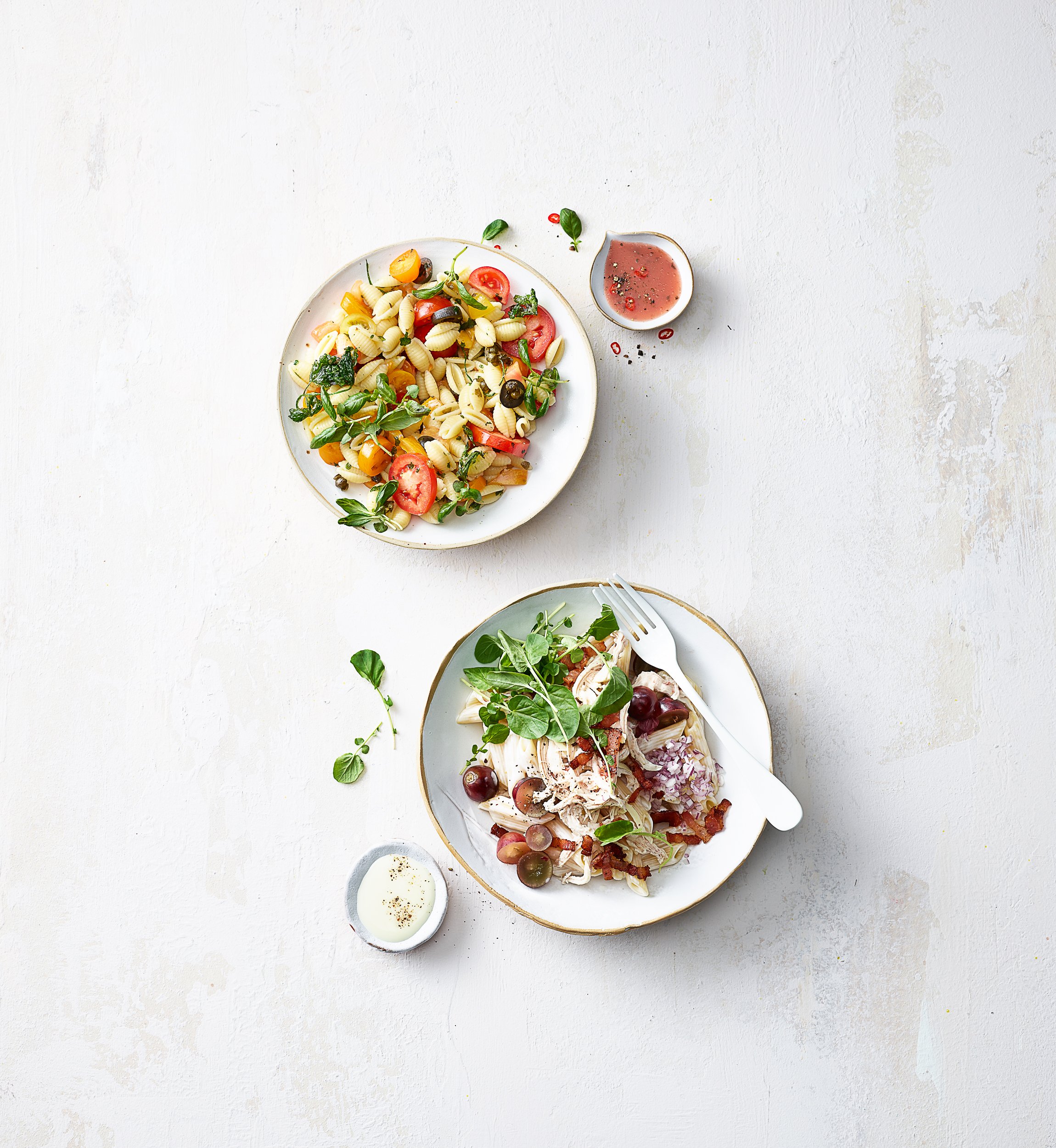 Zwei Teller mit frischen Gerichten auf weißem Hintergrund. Der obere Teller enthält einen Salat aus Gnocchi, Tomaten, Oliven und grünen Blättern. Der untere Teller serviert einen Salat mit Hühnchen, Trauben, Zwiebeln und Blattgemüse. Kleine Schalen m