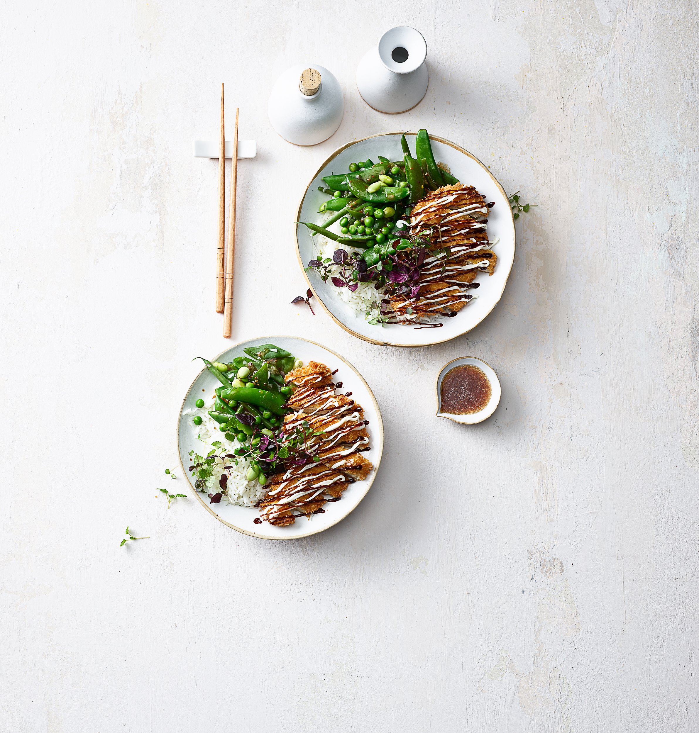 Zwei Teller mit asiatisch inspiriertem Essen, darunter gebratenes Fleisch mit Sauce und grünes Gemüse. Daneben sind Essstäbchen, kleine Keramikbehälter und ein kleiner Becher mit Sauce. Minimalistisches Design auf hellem Hintergrund.