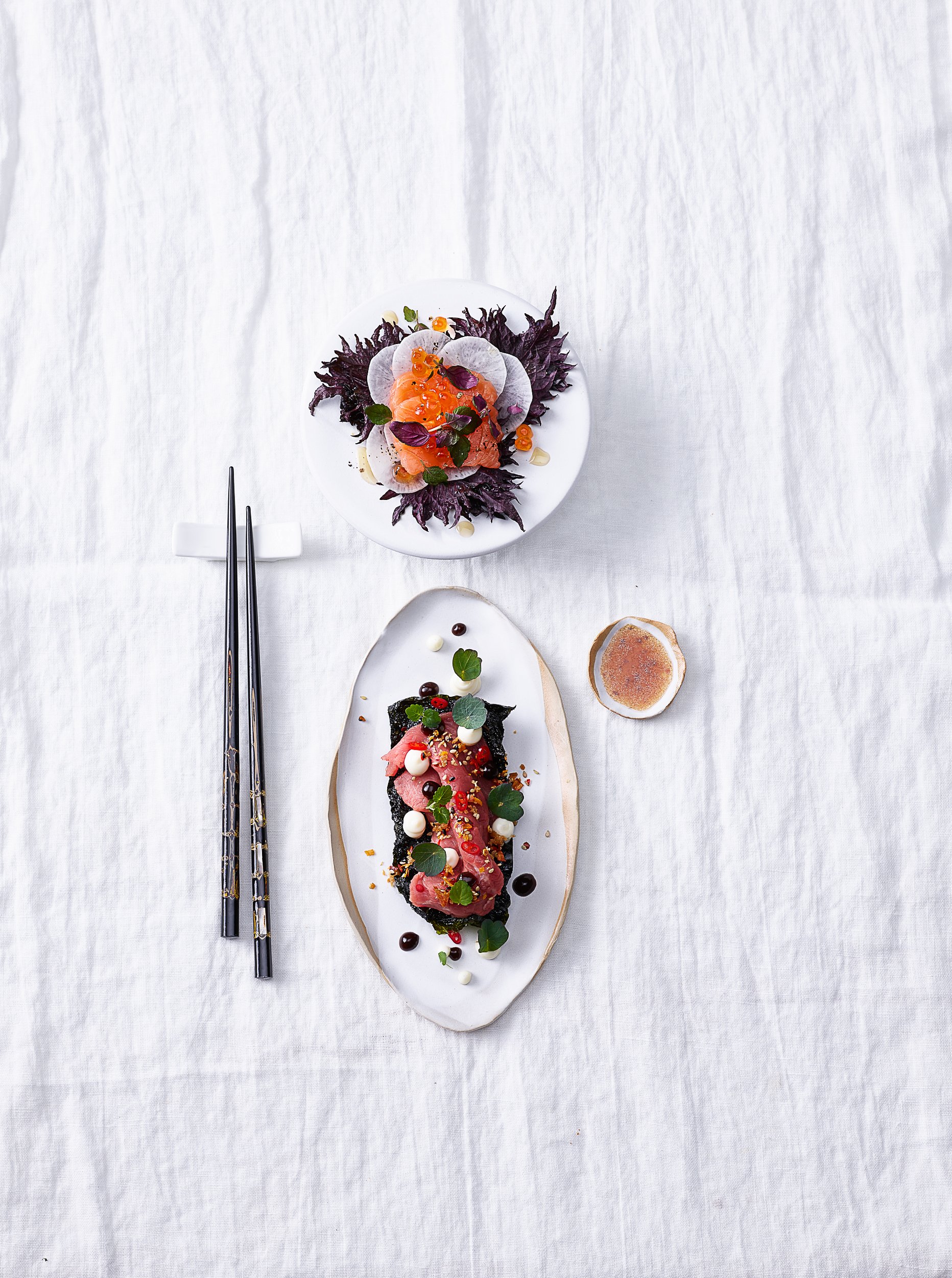 Japanische Gourmetgerichte auf weißen Tellern, dekoriert mit frischen Kräutern und Fisch, neben Essstäbchen auf einer weißen Tischdecke.