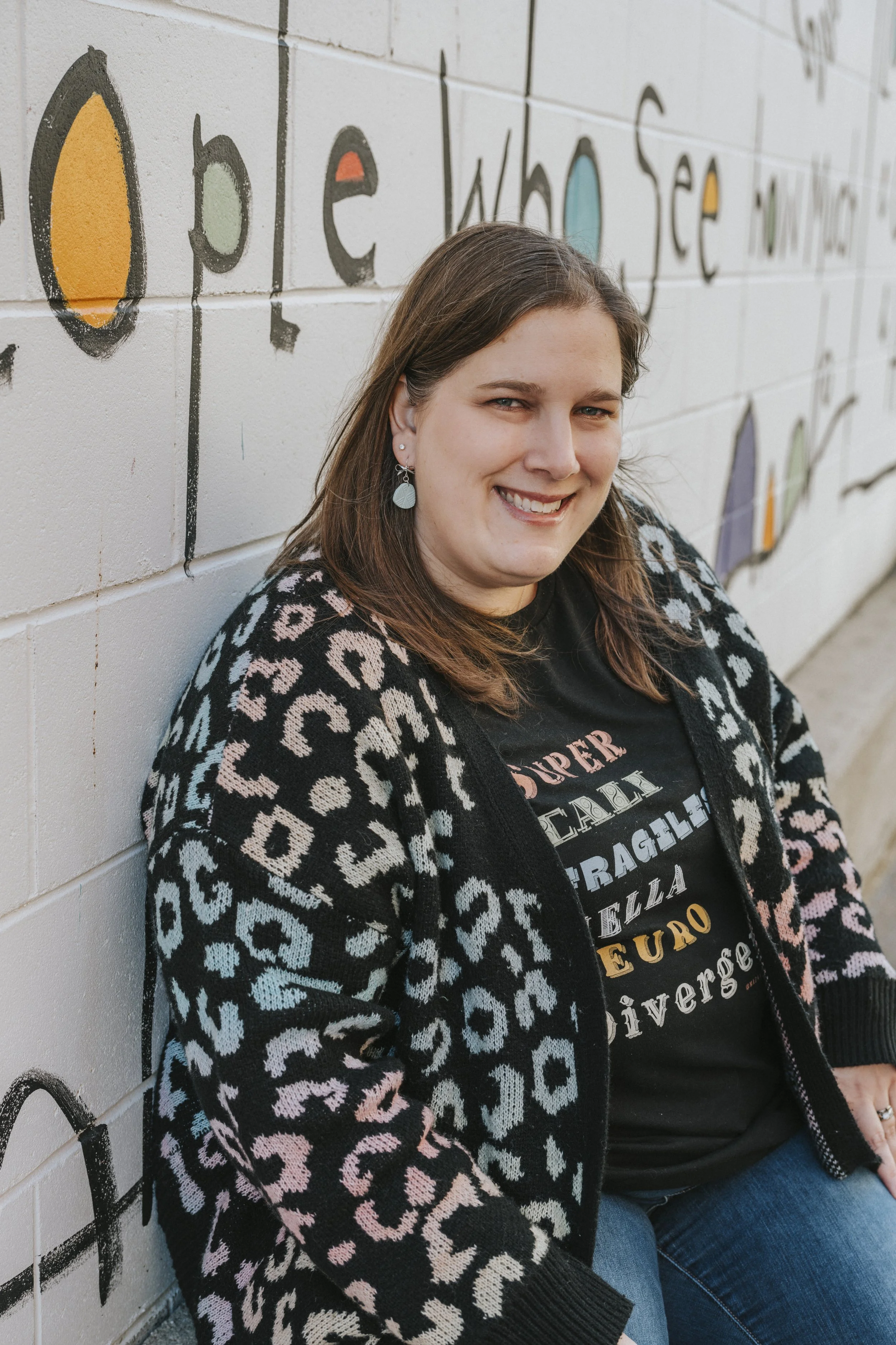 Photo of Bobbi-Jo Molokken, therapist, wearing black t shirt with lettering and black cheeta print sweater with a StoryPeople mural in the background.