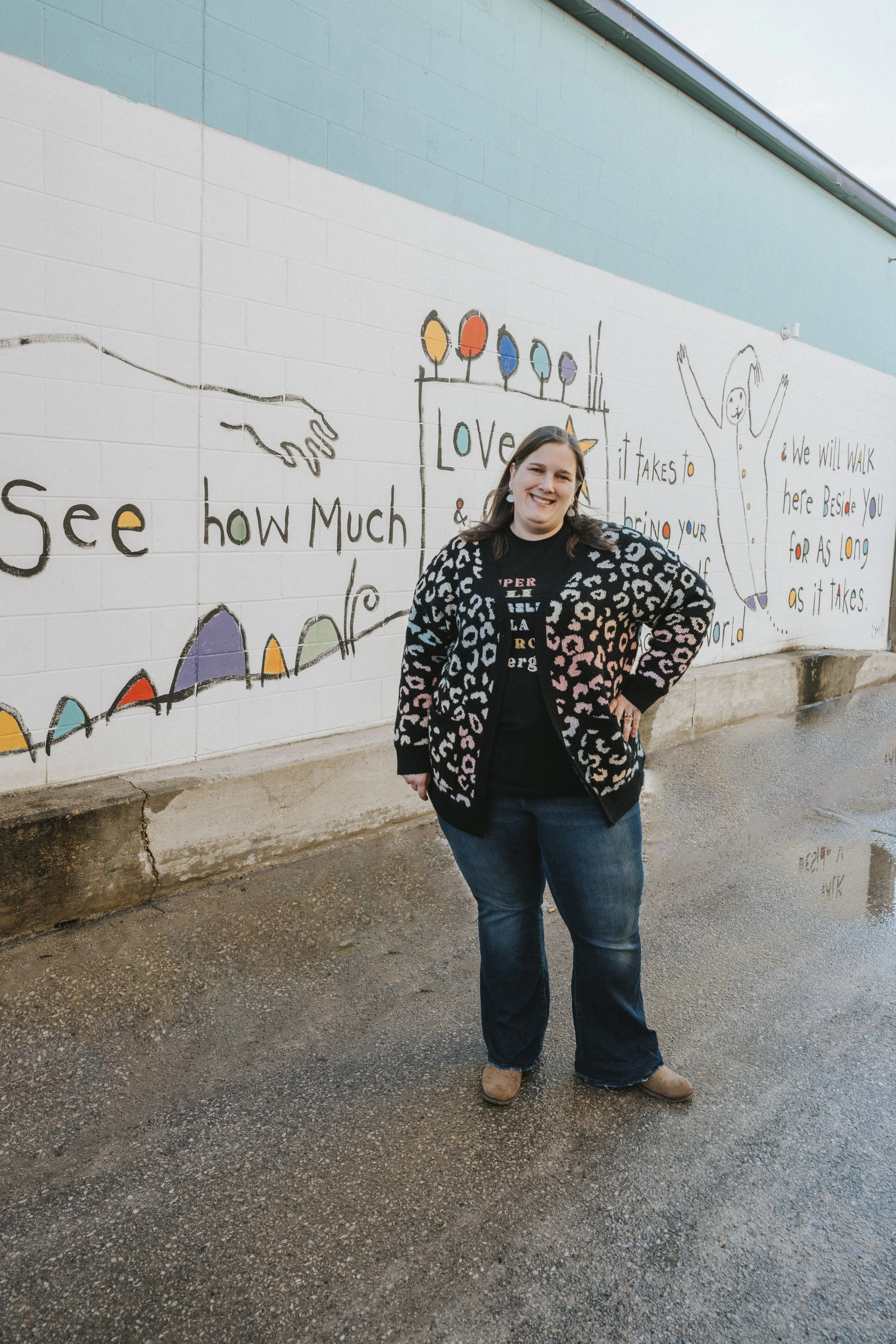 Therapist Bobbi-Jo Molokken stands in front of Storypeople mural in downtown Decorah Iowa.  She is wearing a black sweater with rainbow cheetah print, black tshirt with wording and jeans.