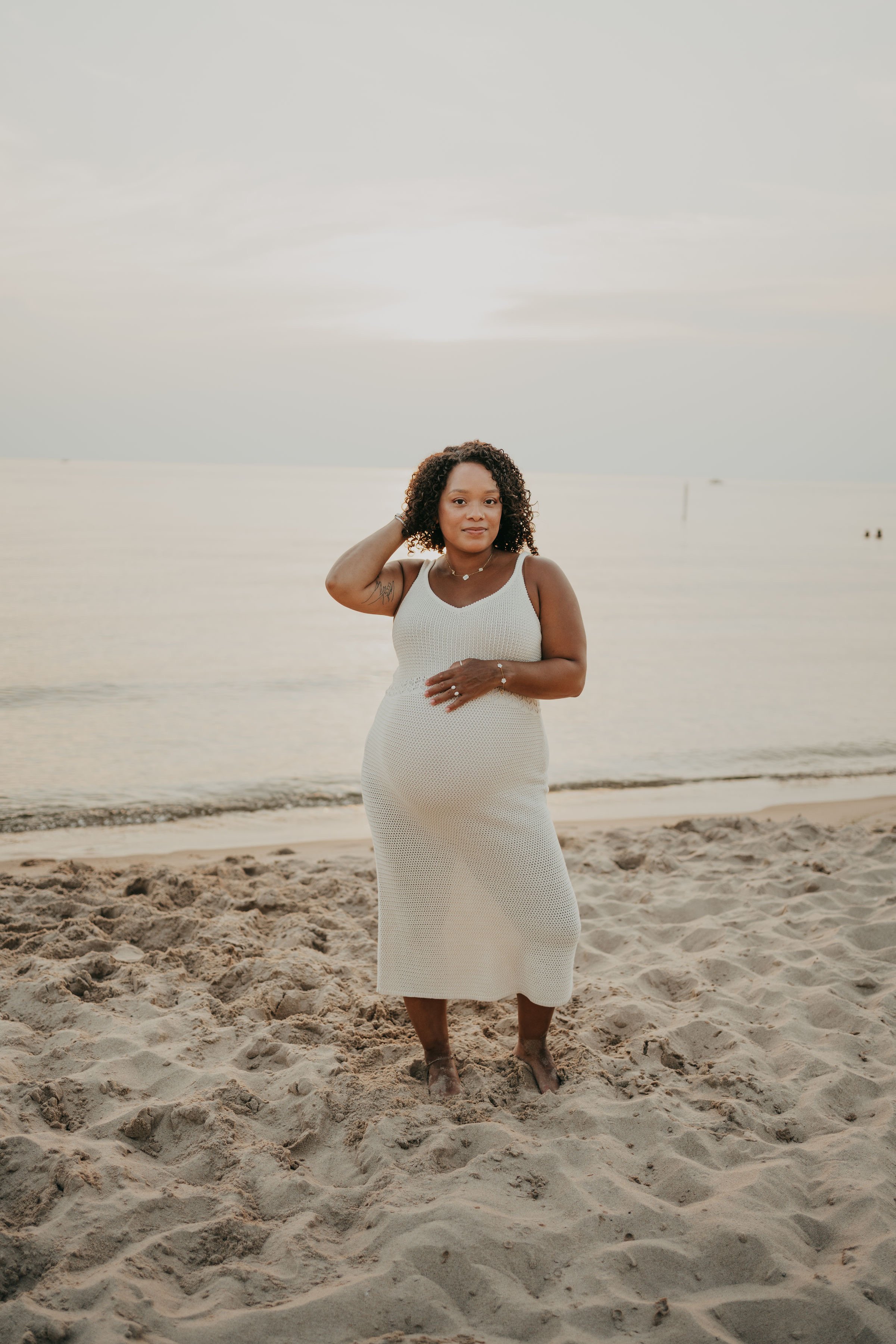 A woman standing on a sandy beach near the water, wearing a white sleeveless dress, with curly hair, jewelry, and bare feet, during sunset or sunrise.