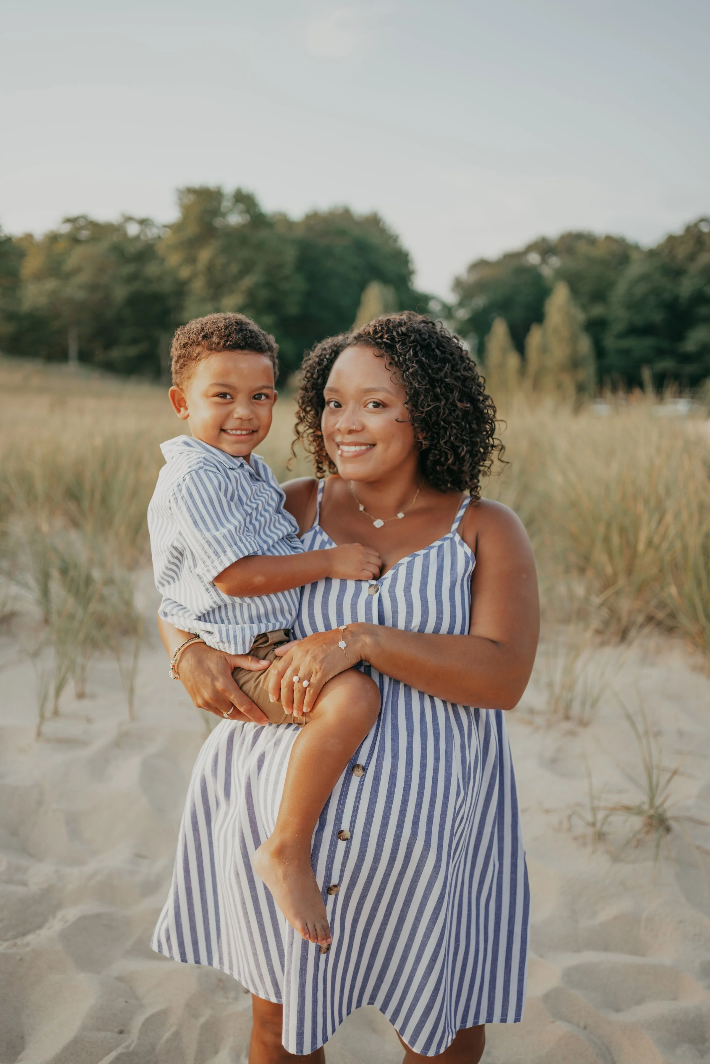 A smiling woman with curly hair holding a young boy with short curly hair at the beach. They are both wearing blue and white striped outfits. The background features sand, grass, trees, and a partly cloudy sky.