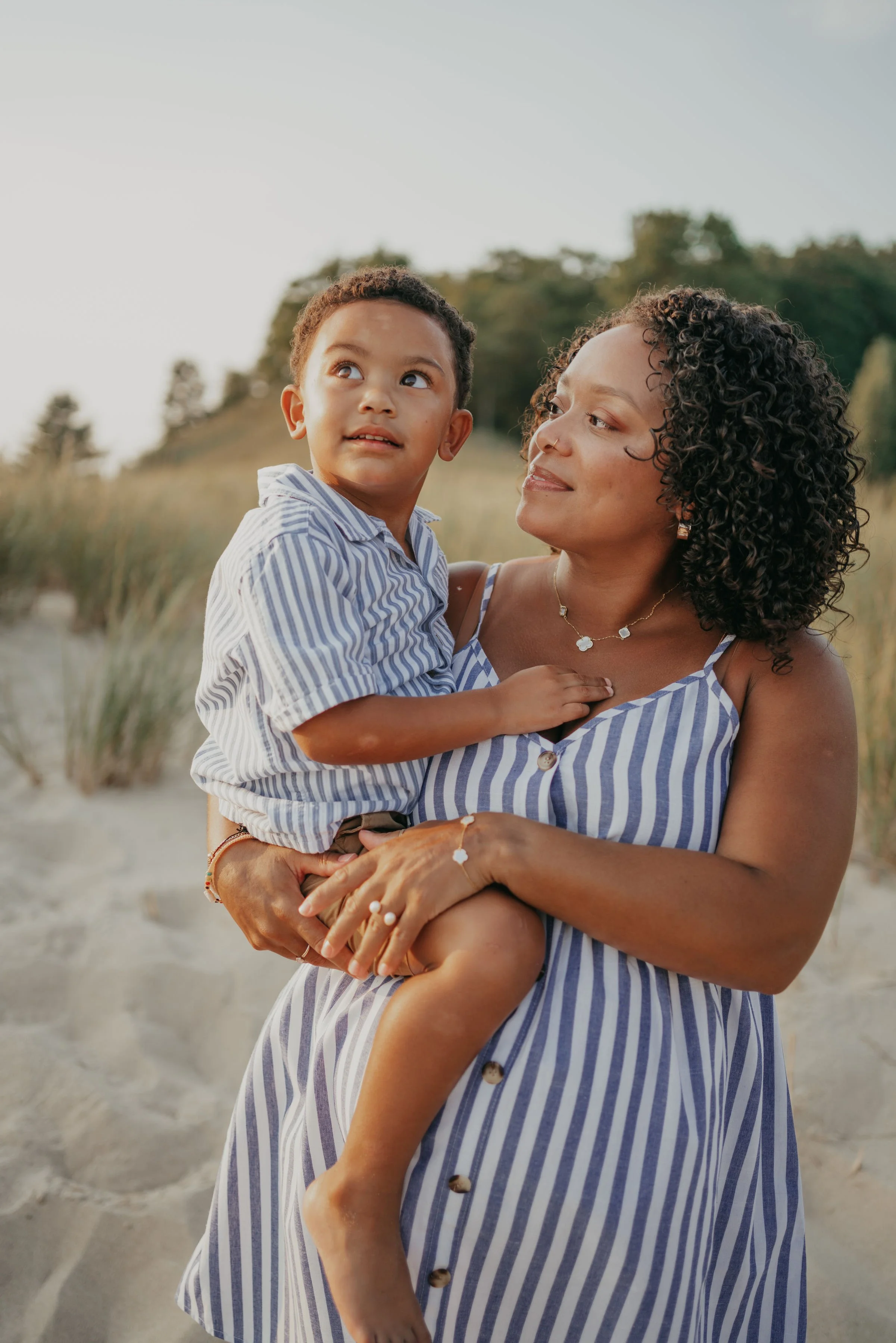 A woman with curly hair and wearing a striped dress holds a young boy with short curly hair and a striped shirt outdoors, with trees and a clear sky in the background.