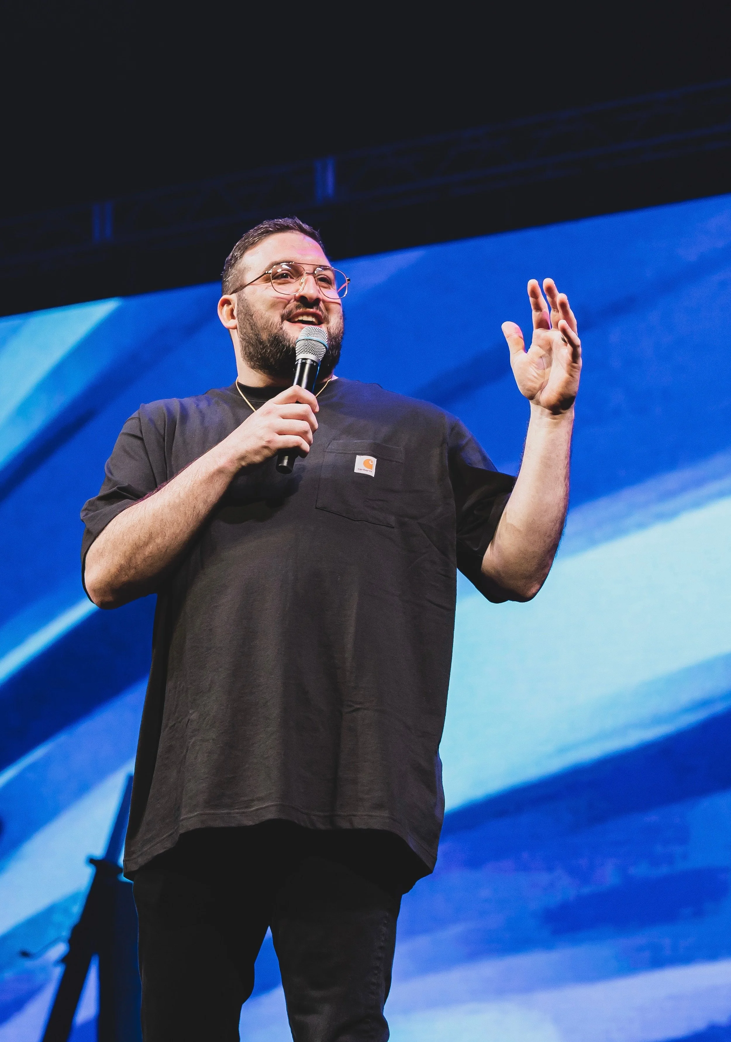 A man with glasses and a beard speaking into a microphone on stage against a blue abstract background.