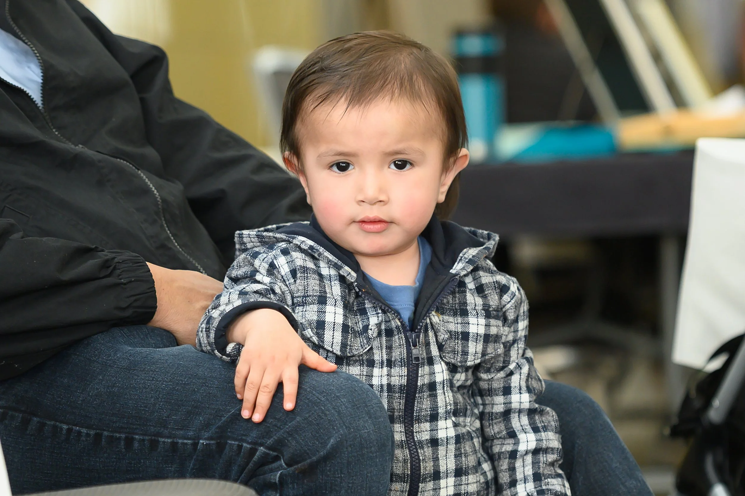 A young boy with light skin and brown hair wearing a checkered jacket, sitting indoors with a serious expression.