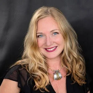 A smiling woman with long blonde hair, blue eyes, wearing a black top and a necklace, against a dark background.