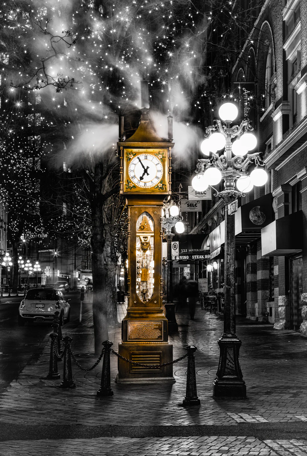 A vintage street clock on a city sidewalk at night, with steam rising from the top and sparkling holiday lights in the background. Street lamps illuminate the scene, with buildings, parked cars, and pedestrians visible.