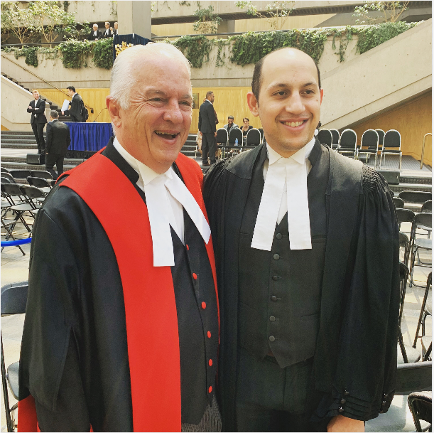 Two men in academic robes at a graduation ceremony, smiling and posing for a photo indoors.