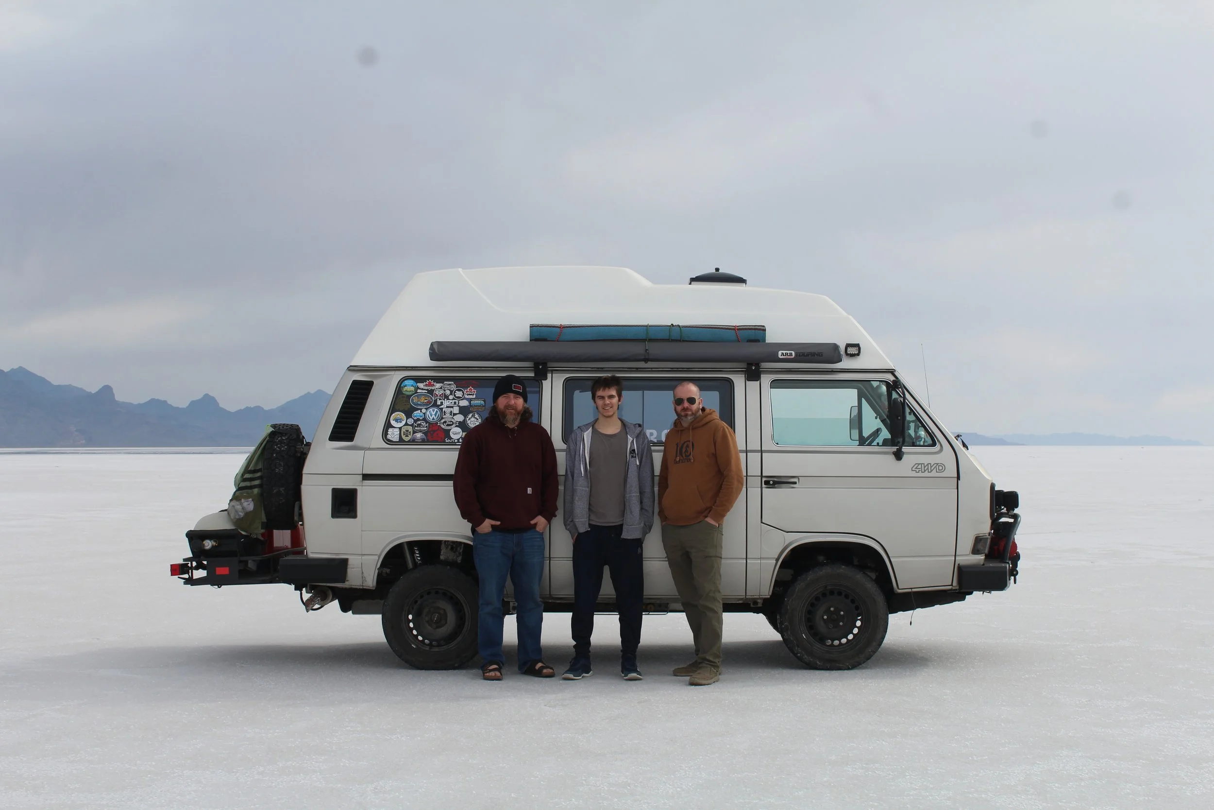 Chris Moxon, Austin Campbell and Ryan Campbell are standing in front of a white camper bus parked on a vast salt flat. The sky is clear, and distant mountains are visible. The bus features a pop-up roof, and a spare tire mounted on the back.