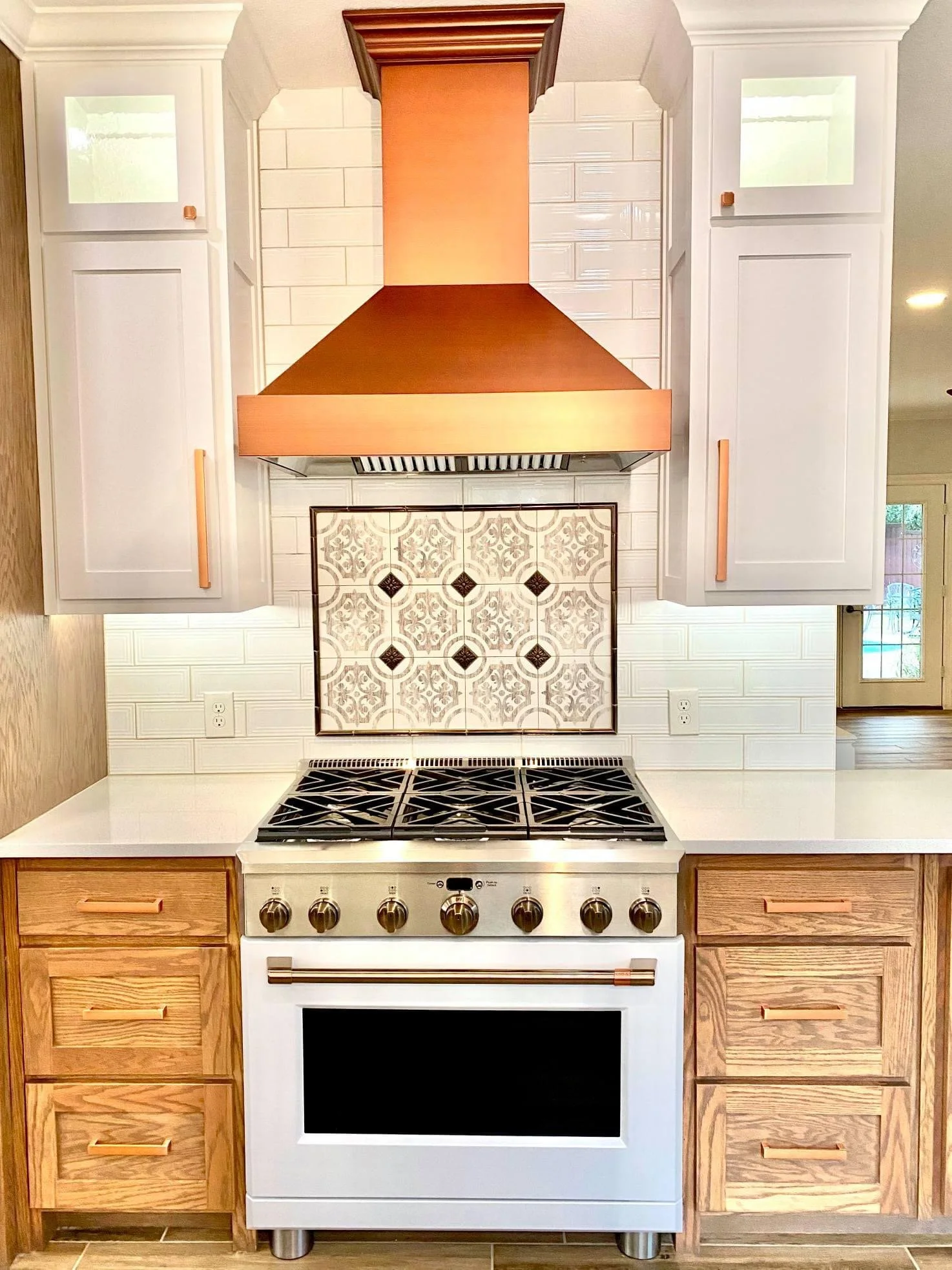 A modern kitchen stove with a white oven, black cooktop, and bronze knobs, flanked by wooden cabinets on each side, with a decorative tiled backsplash and white upper cabinets, and a window to the right.