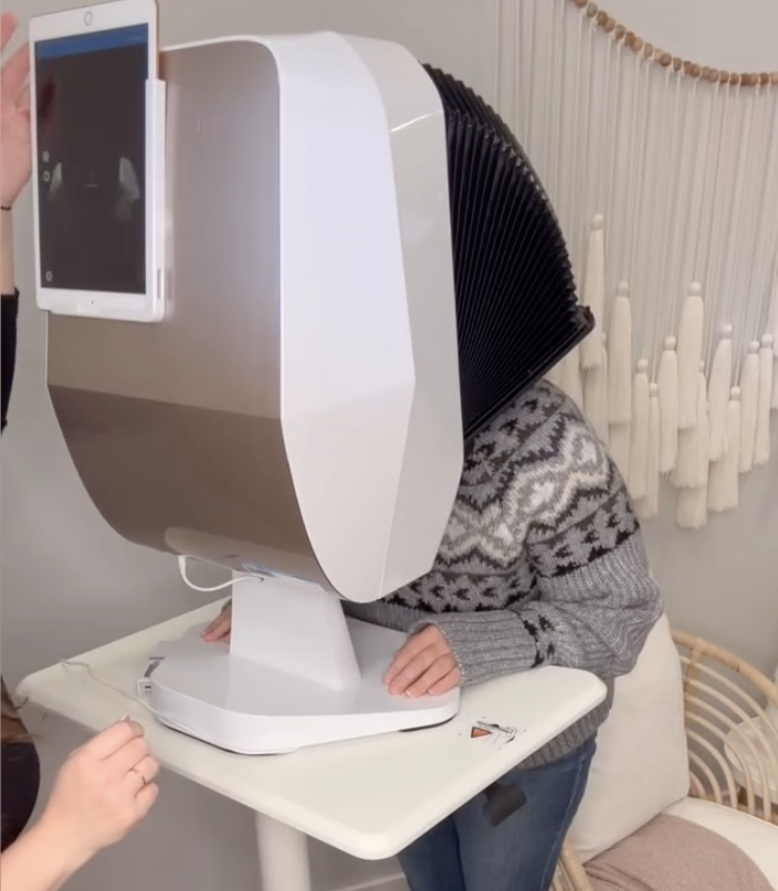 A person with their head inside a large, white, cube-shaped machine with a screen on one side, sitting at a small white table.