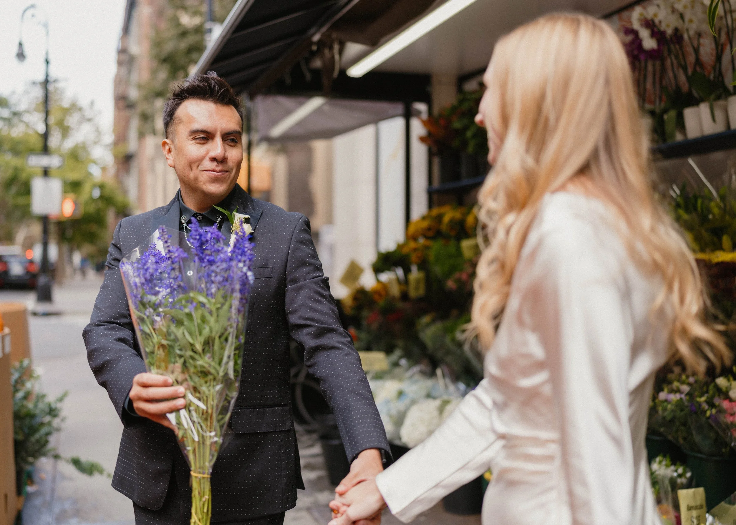 A man in a black suit and a woman in a white jacket holding hands in front of a flower stand on a city street. The man is holding a bouquet of purple flowers.