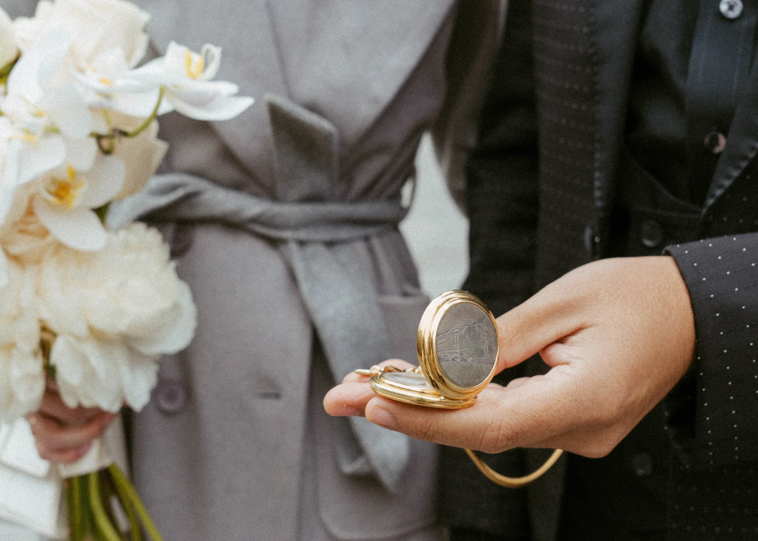 A person holding a pocket watch with gold trim, while another person holds a bouquet of white flowers, possibly orchids, in the background. The person with the pocket watch is wearing a black shirt with white polka dots, and the person with the bouqu