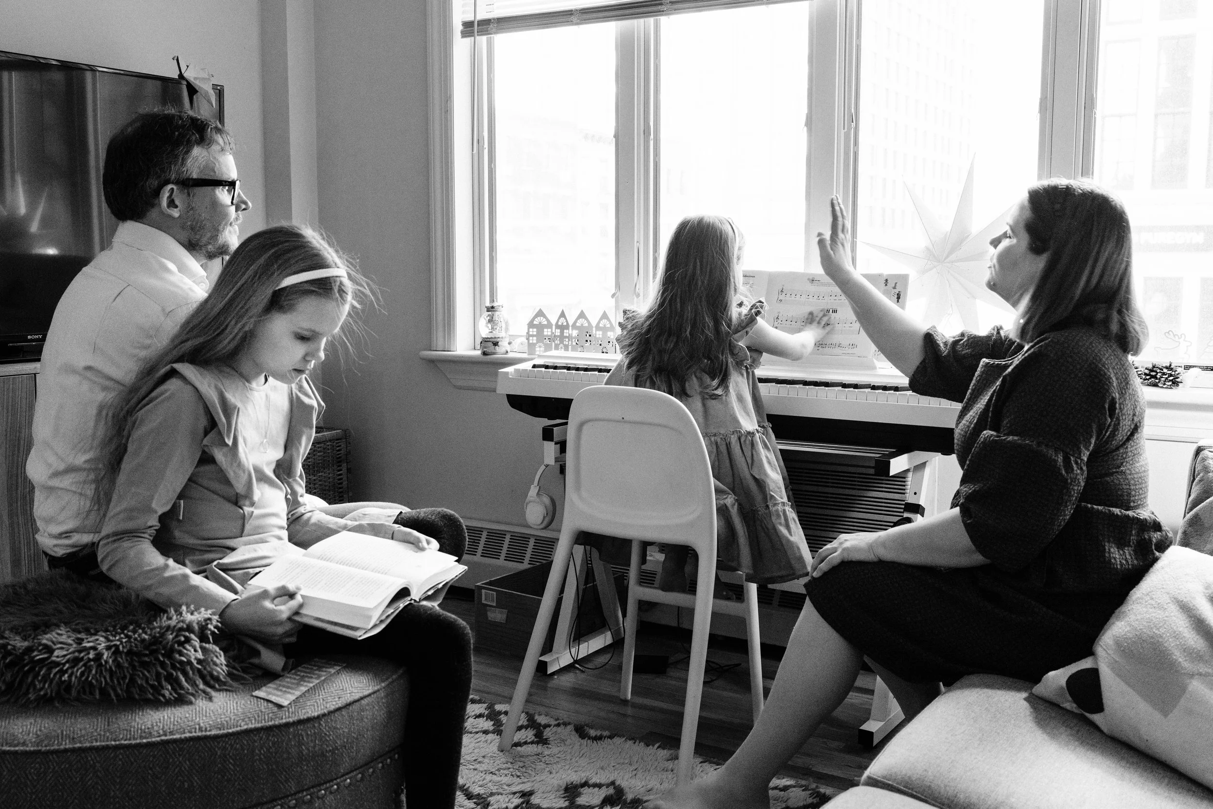 A family in a living room with two children and two adults, one girl playing a keyboard and another reading a book, with adults engaging with them.
