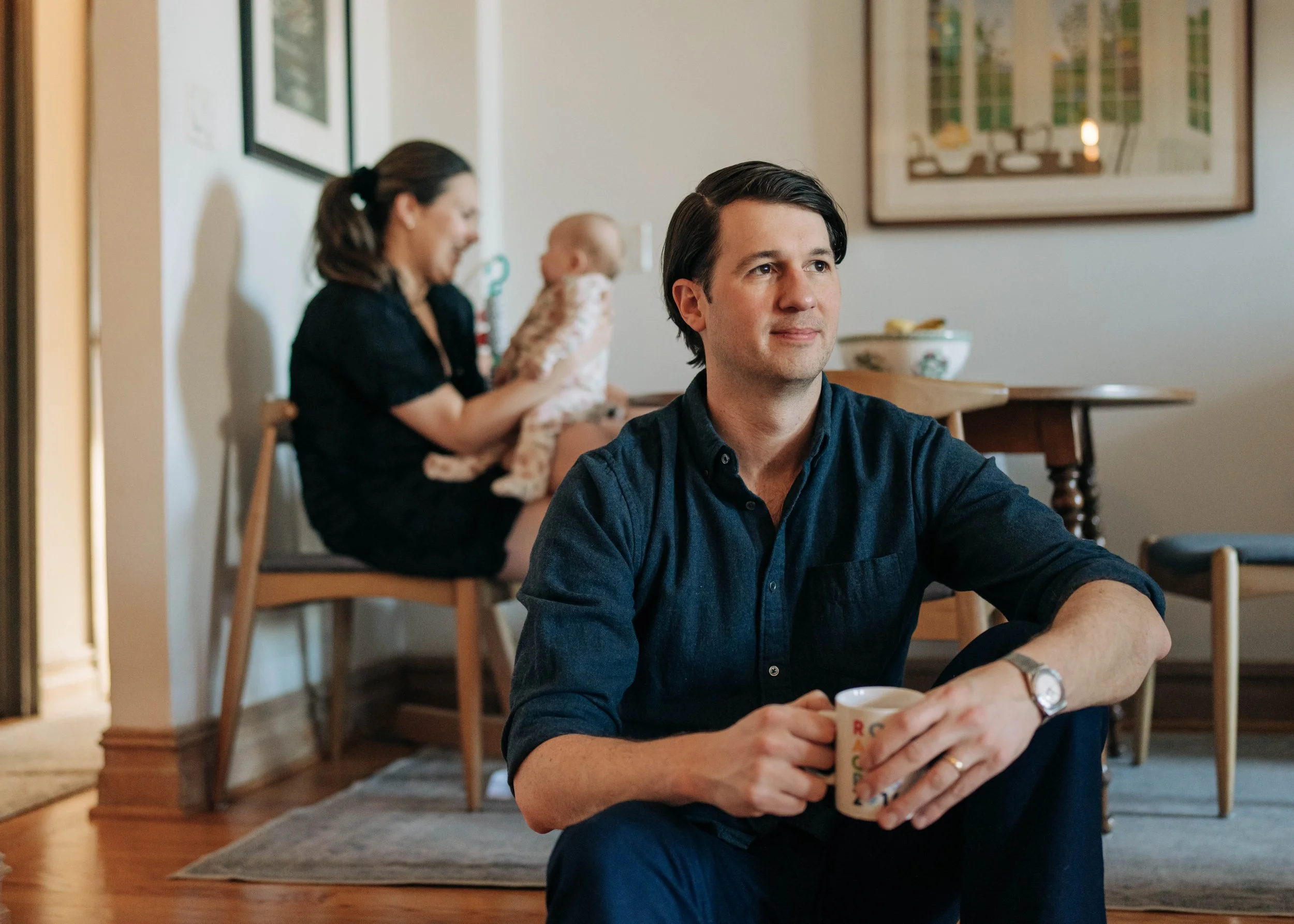 A man sitting on the floor holding a coffee mug, with a woman in the background holding a baby in a dining area of a home.