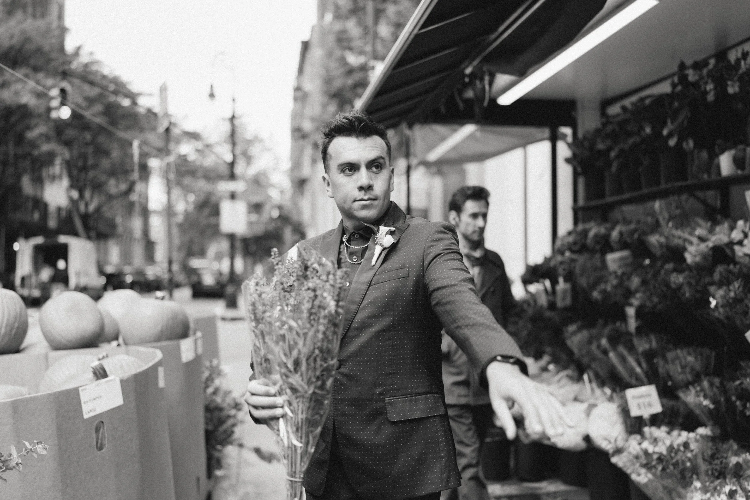 A man in a suit at a street market holding a bouquet of flowers, with another man in the background shopping at a flower stand.