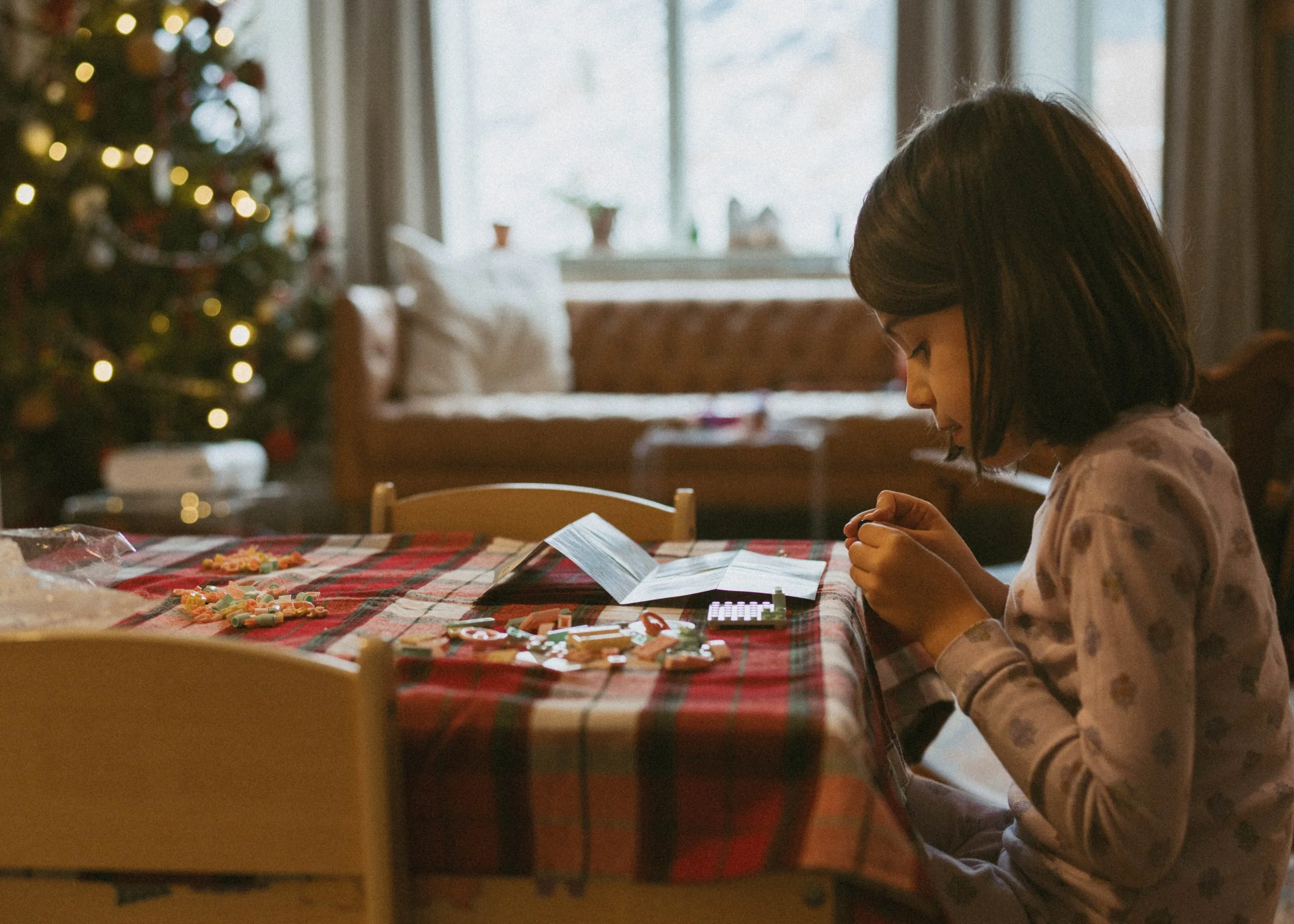 A young girl sitting at a dining table with Christmas candies, a manual, and a calculator, with a Christmas tree in the background.