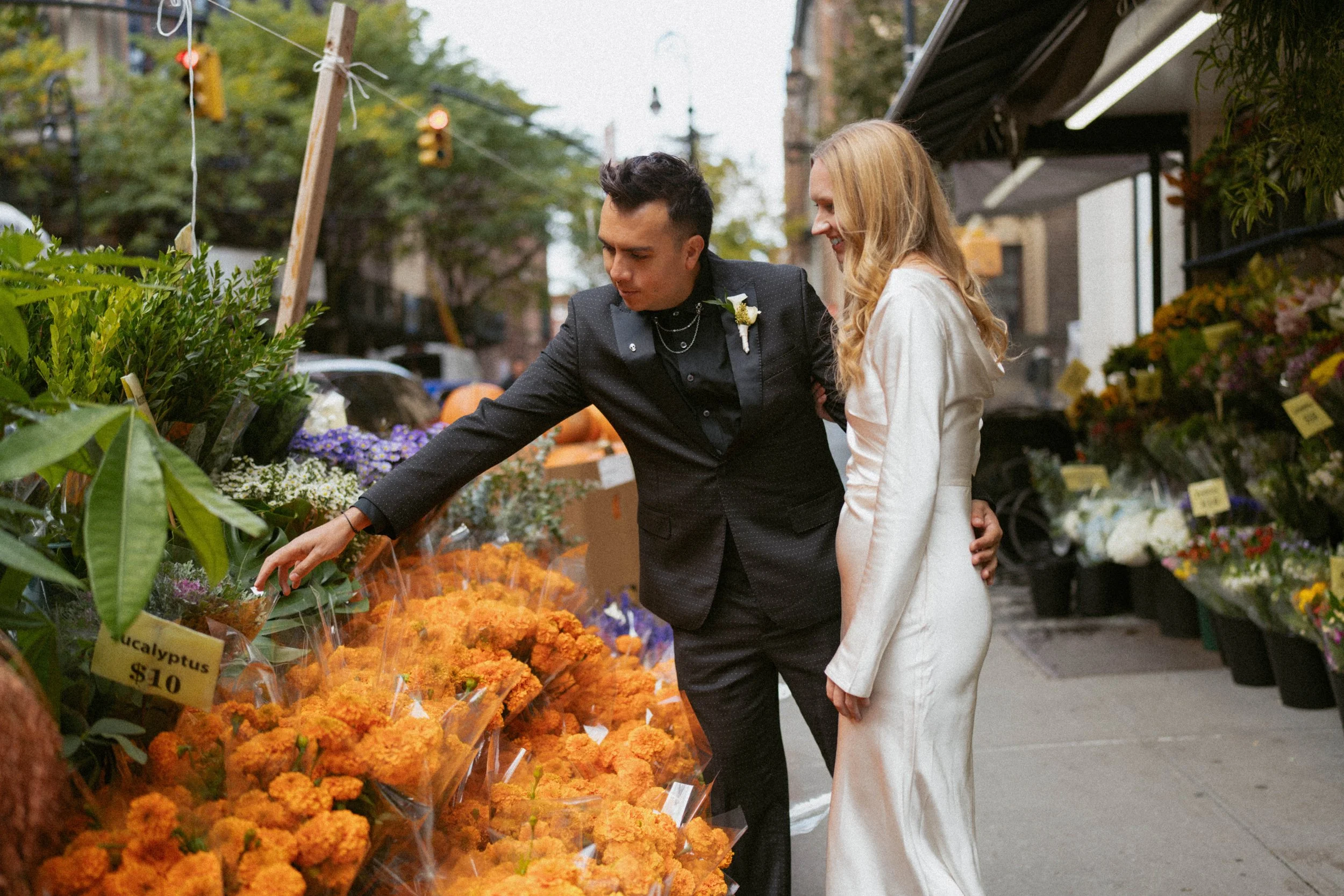 A couple in wedding attire shopping at an outdoor flower market. The groom is selecting orange flowers, while the bride watches with a smile.