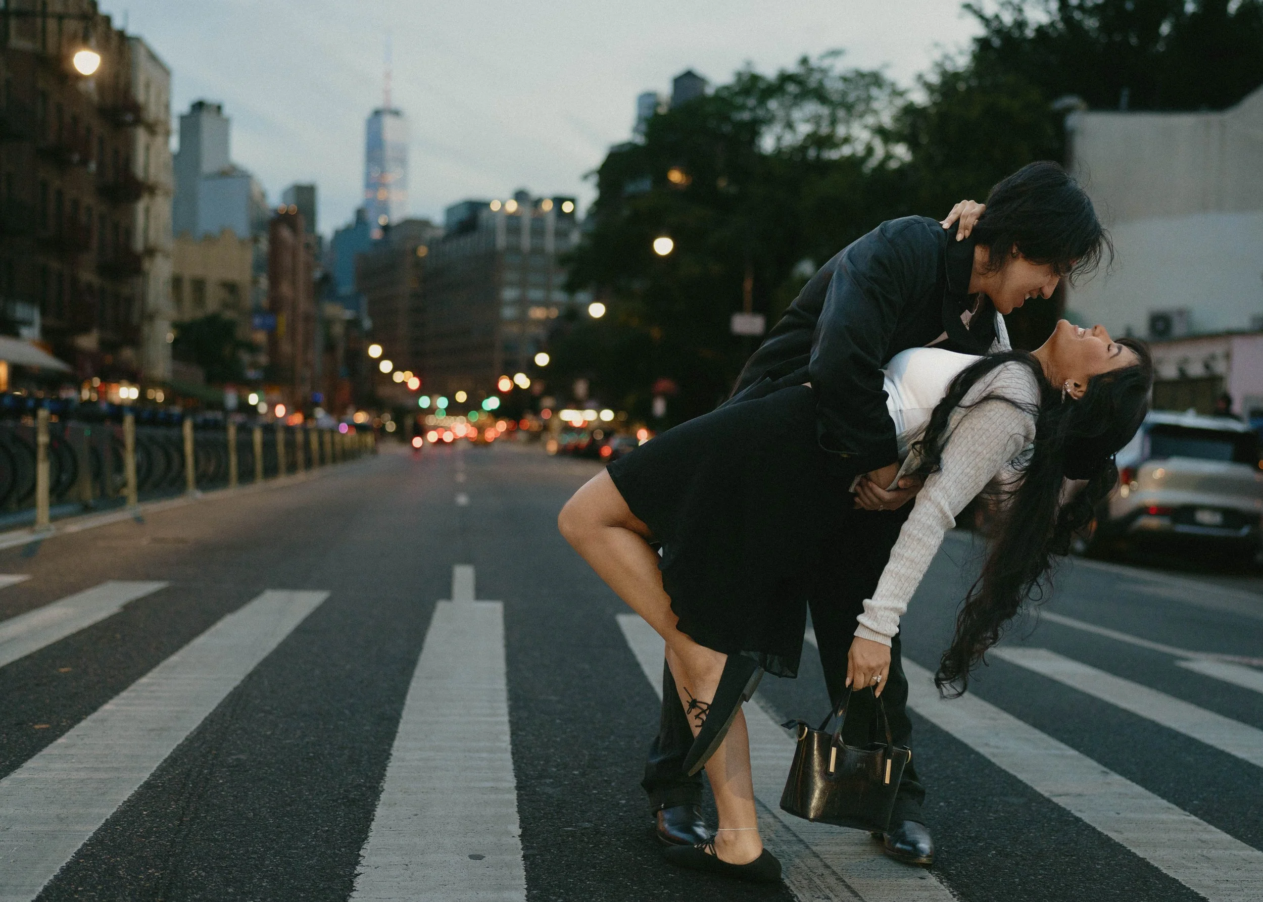 Two women share a joyful moment on a city street during dusk, with one woman leaning over the other in a playful pose, blurred city buildings and lights in the background.