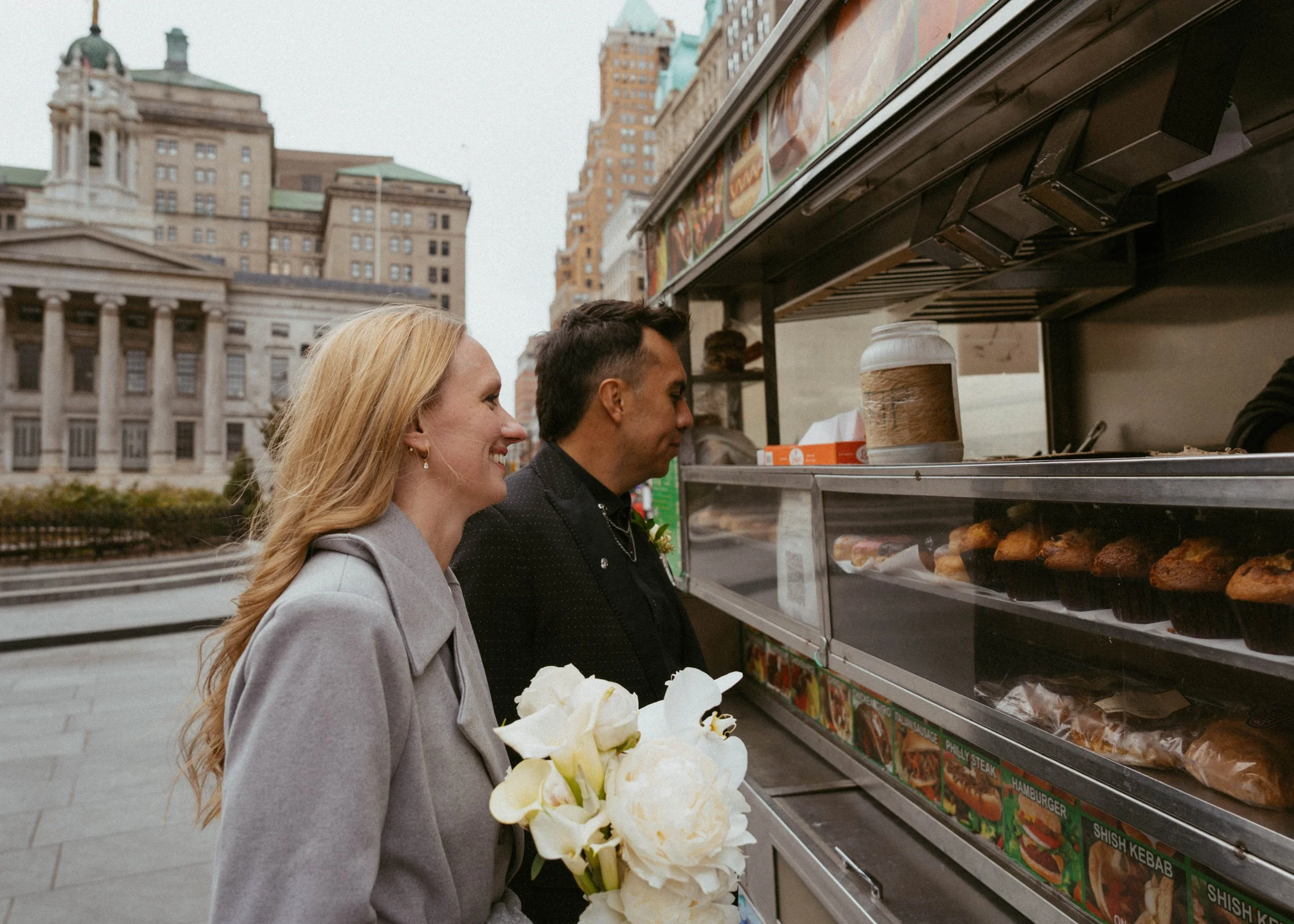 A woman in a gray coat holding a bouquet of white flowers and a man in a black jacket ordering food at a street food stand in an urban area with historic buildings in the background.