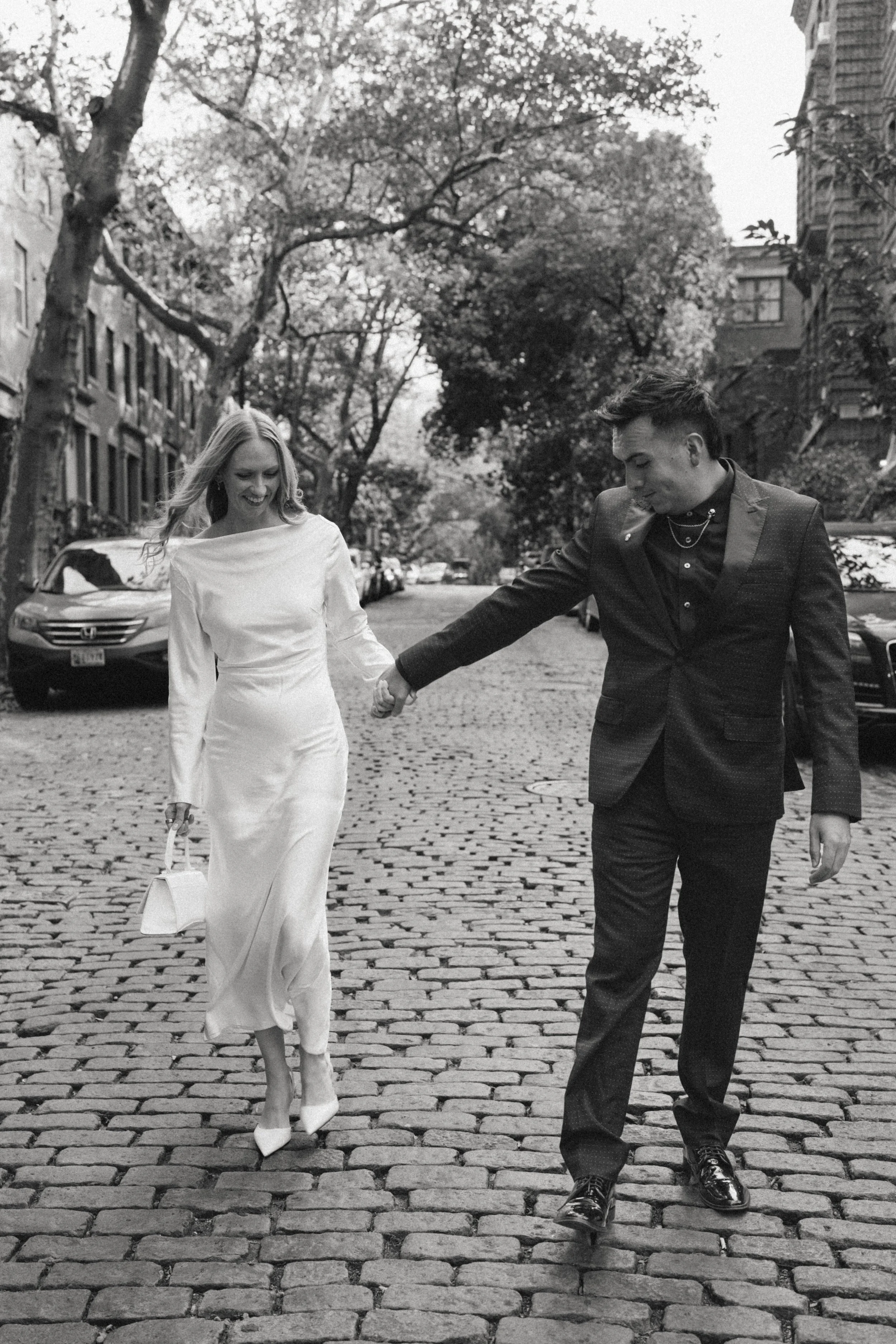 A black-and-white photograph of a couple walking hand in hand on a cobblestone street, with trees and parked cars in the background. The woman is wearing a long white dress and high heels, holding a small handbag, and smiling. The man is dressed in a