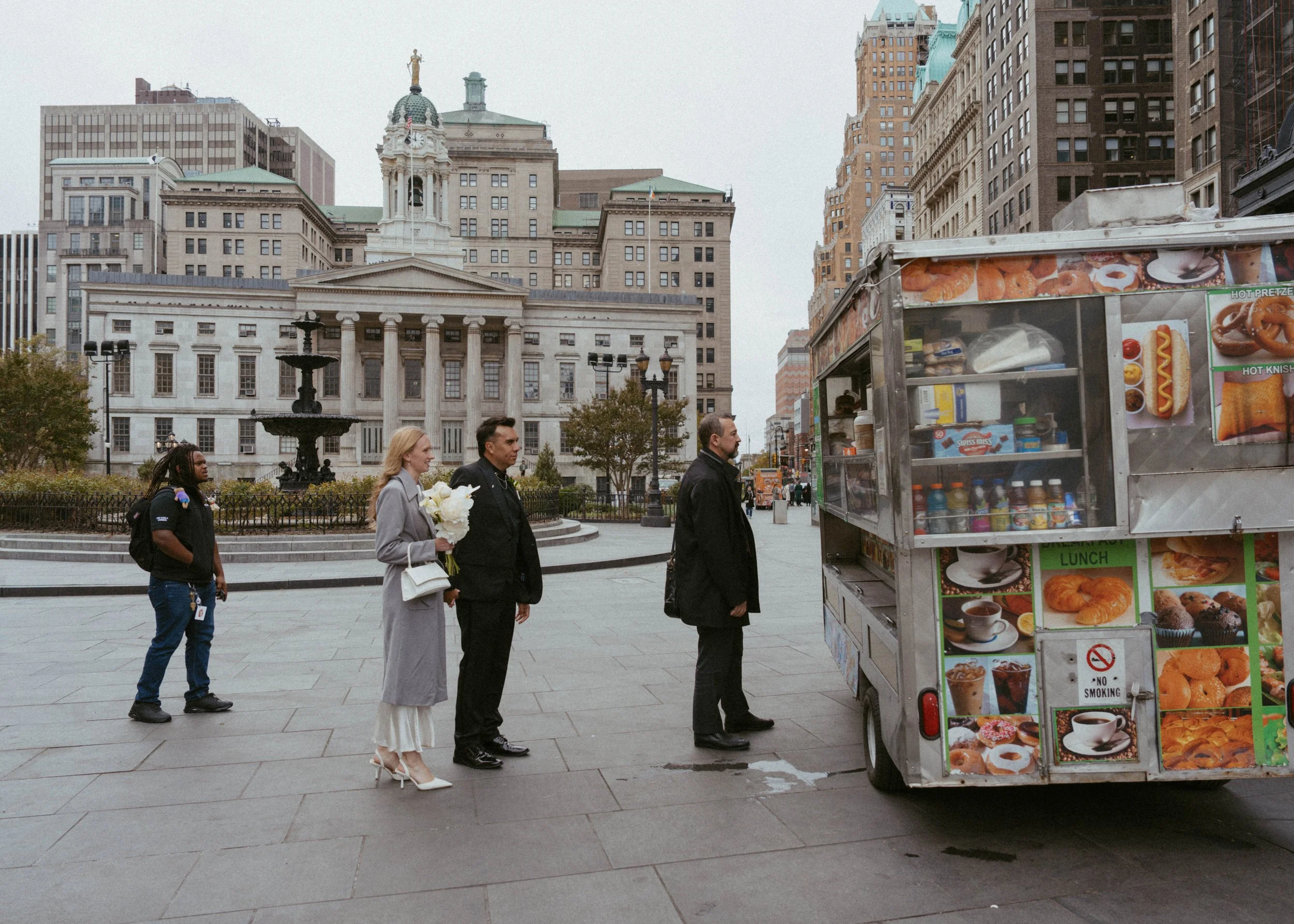 Four people standing in line at a food cart in an urban plaza, with a neoclassical building and skyscrapers in the background.