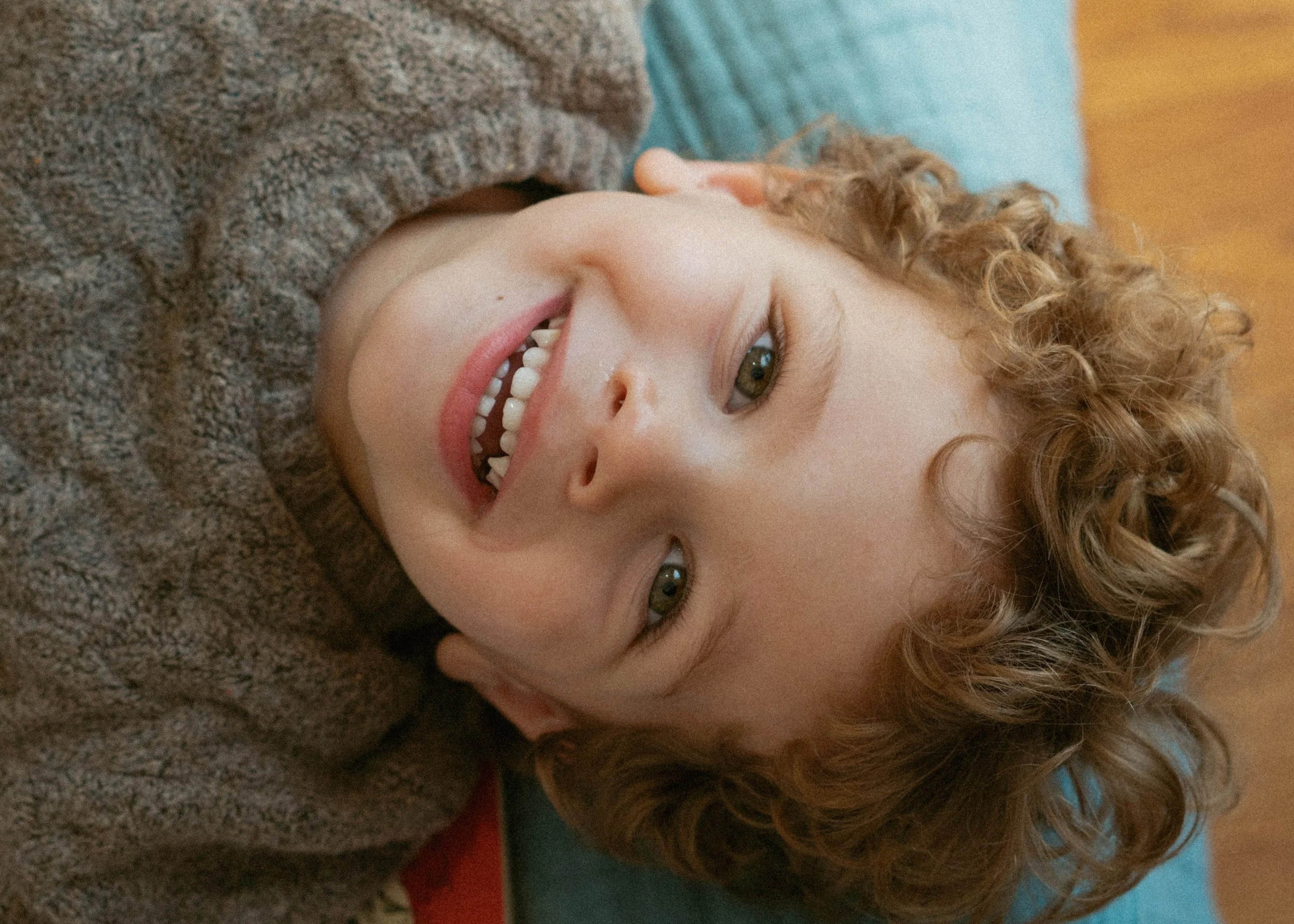 A young boy with curly hair lying on his back, smiling and looking up at the camera.