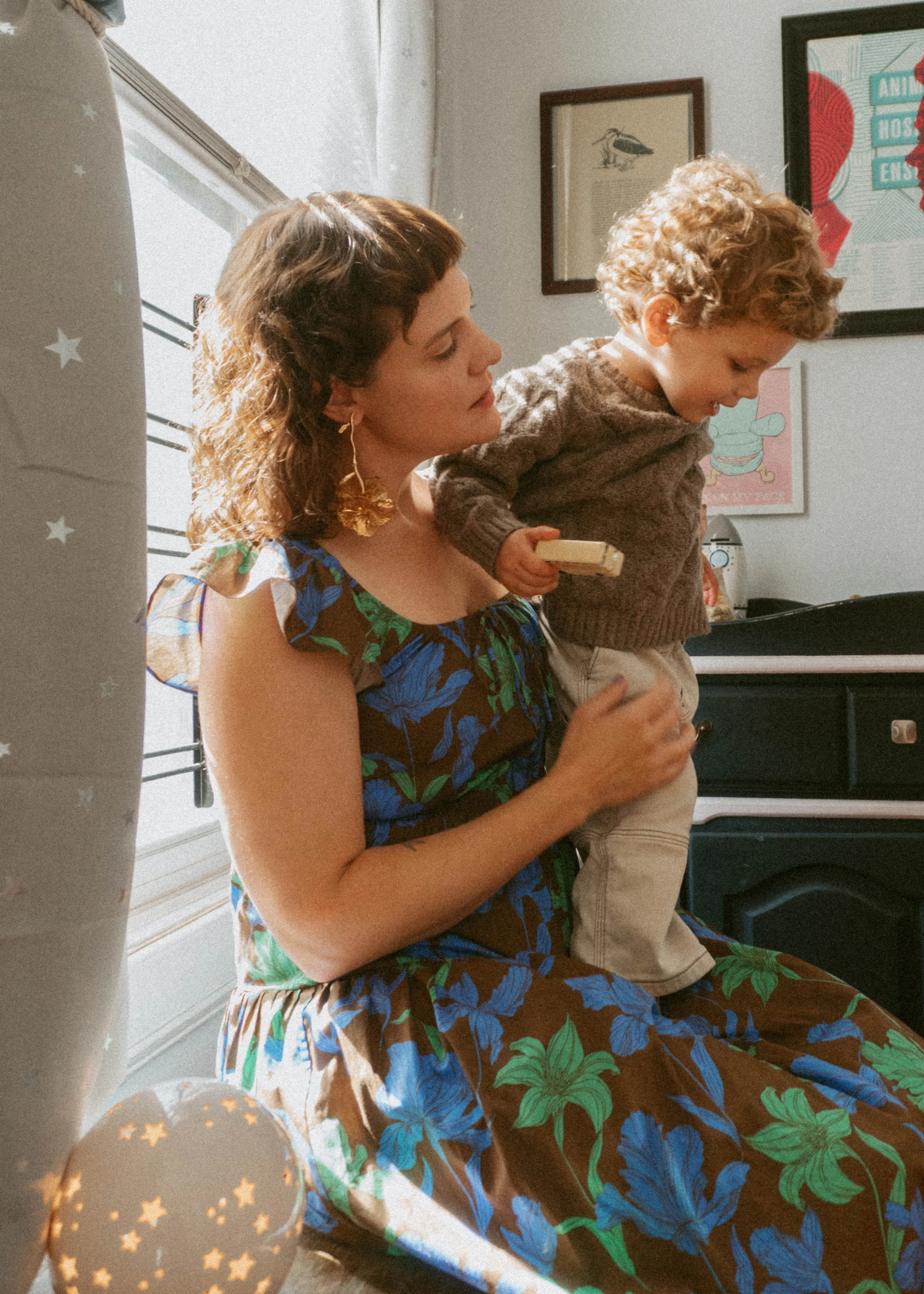 A woman sitting by a window holding a young boy on her lap. The boy is looking down and holding a small object in his hand. There are balloons and framed artwork on the wall behind them.