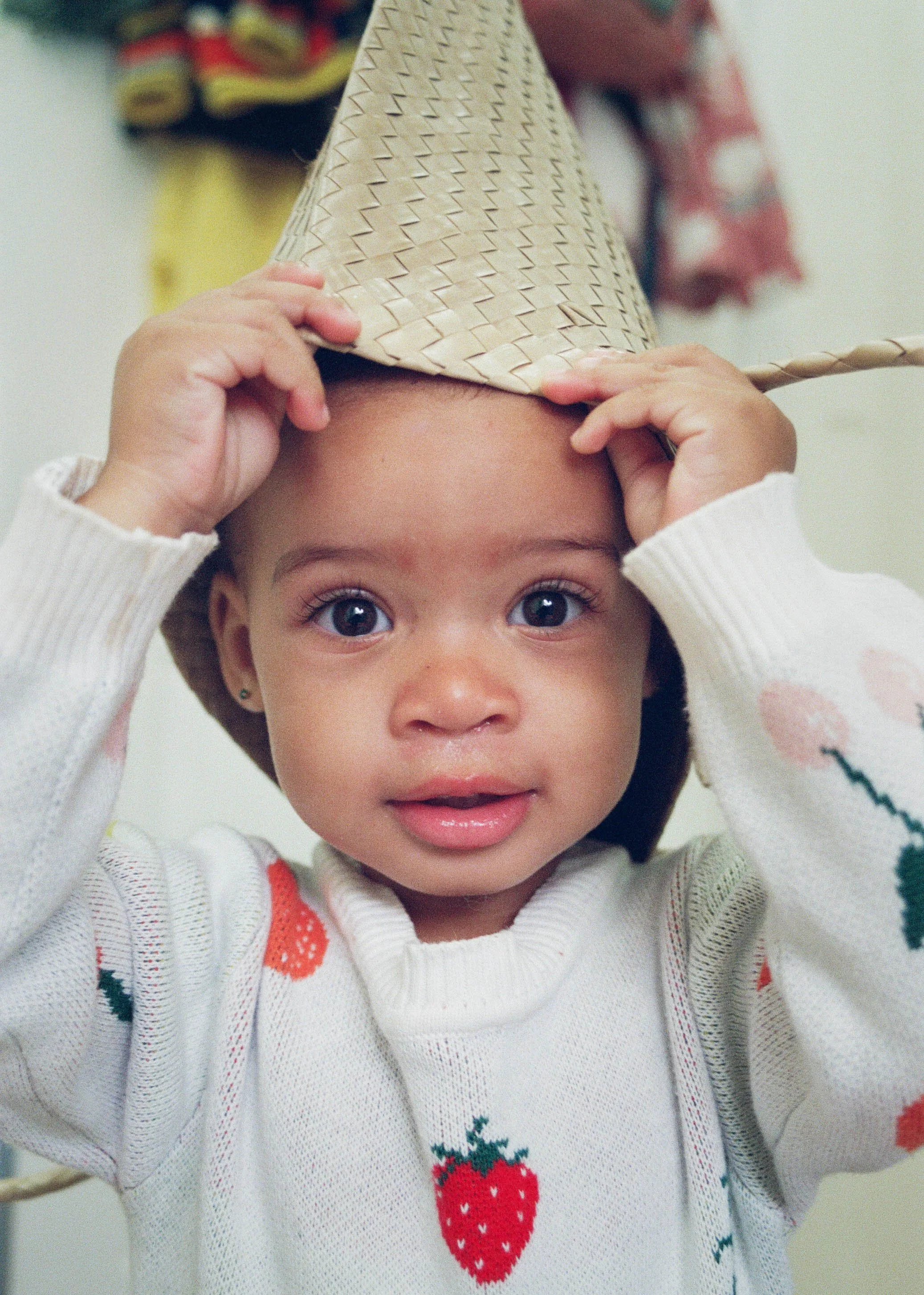 A young girl with big brown eyes and a white sweater with strawberry patterns is putting on a woven straw hat.