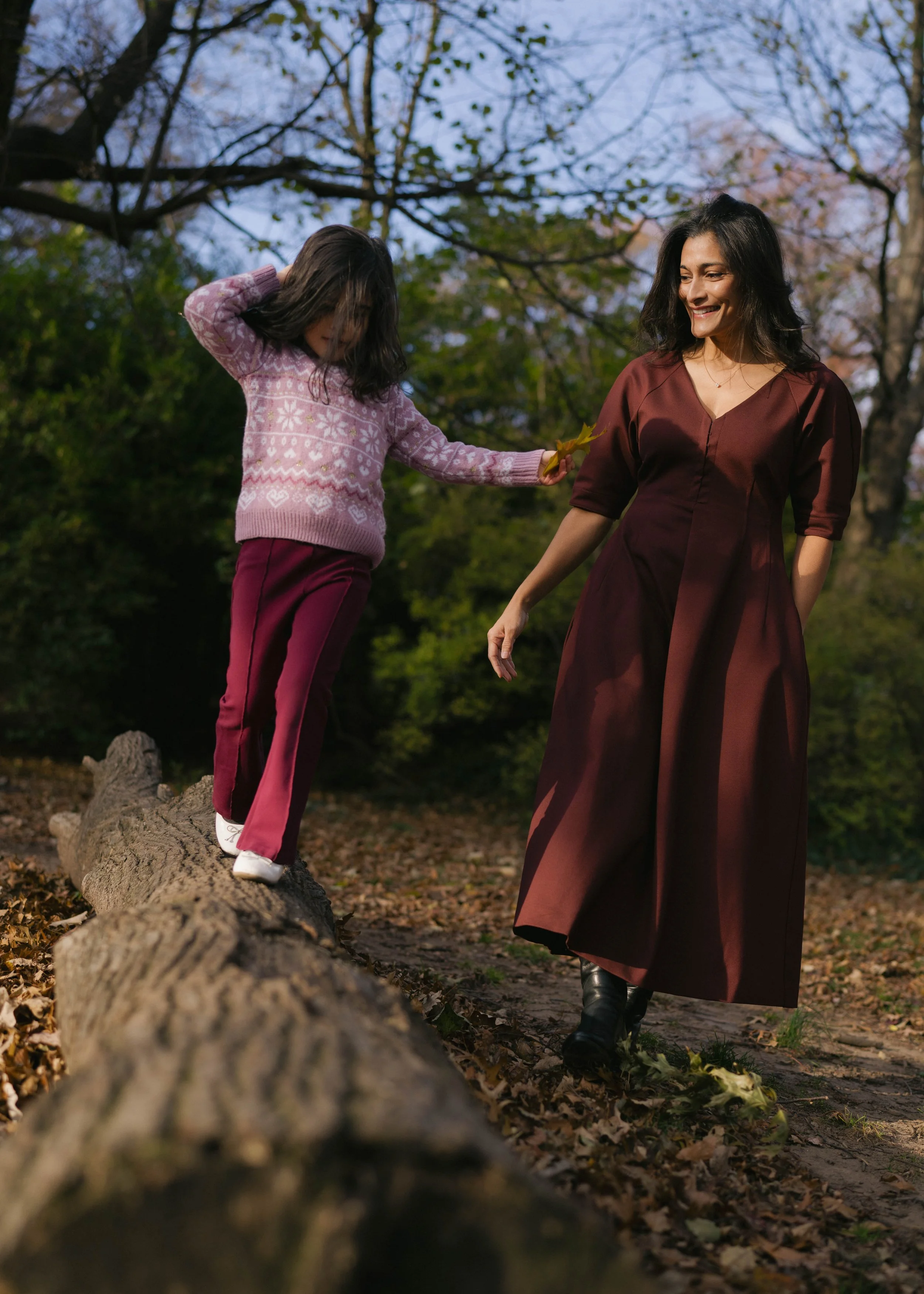 A woman and a girl walking on a fallen tree trunk in a park during fall, with trees and colorful leaves in the background, the woman holding a yellow leaf.