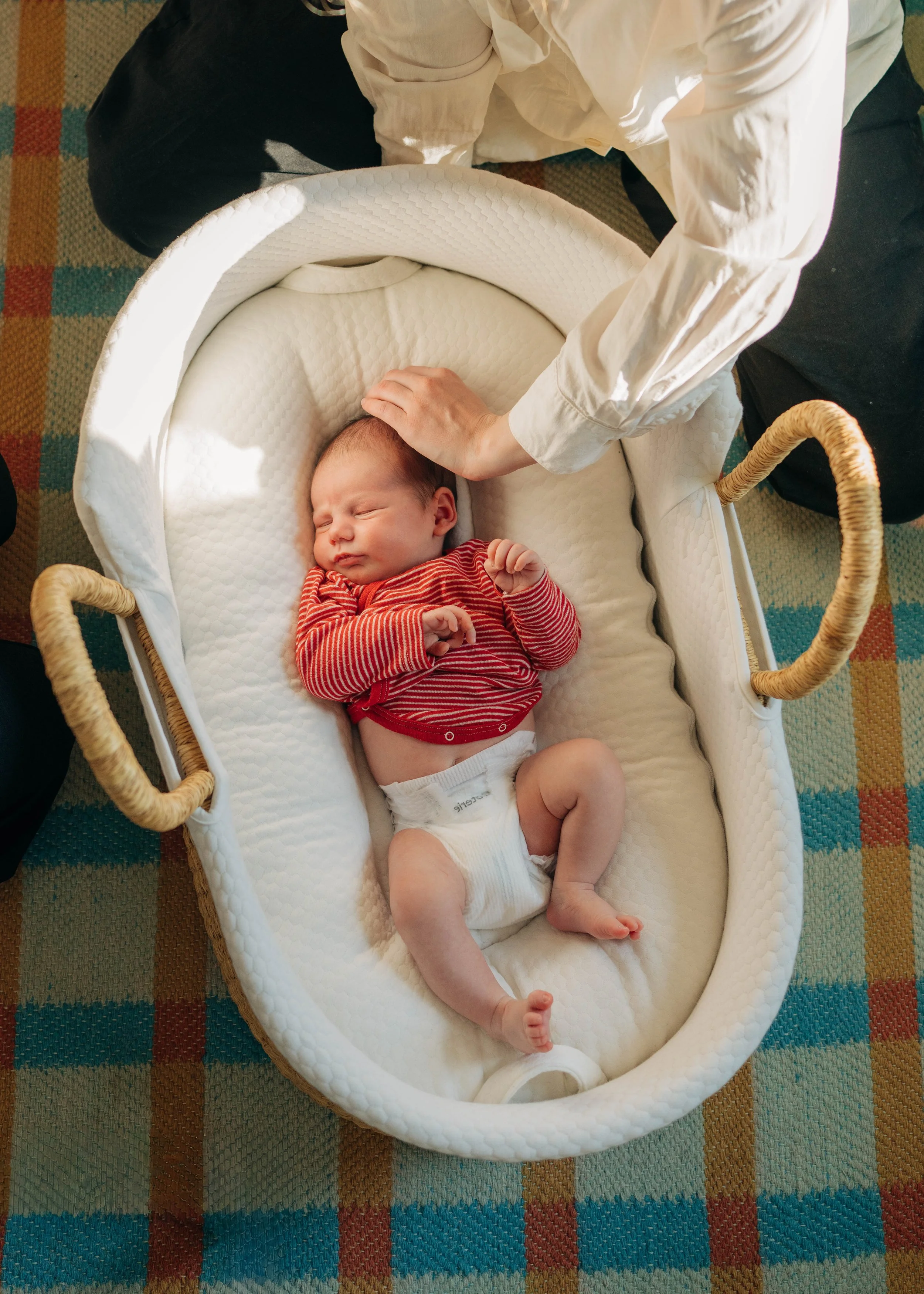 A baby in a white bassinet with a white textured blanket, sleeping peacefully with a red and white striped shirt and a diaper, a person gently touching the baby's head.