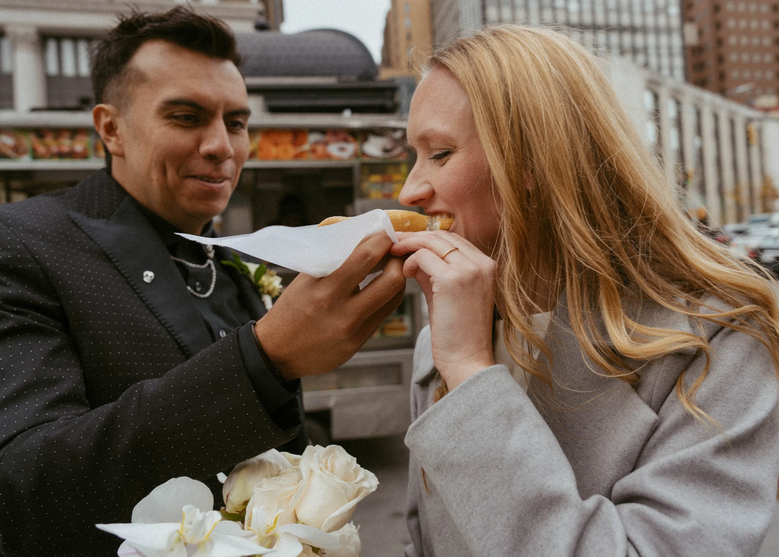 A man dressed in a black suit with a wedding boutonniere is holding a hot dog for a woman with long red hair, who is about to take a bite. They are outside in an urban setting with buildings and cars in the background.