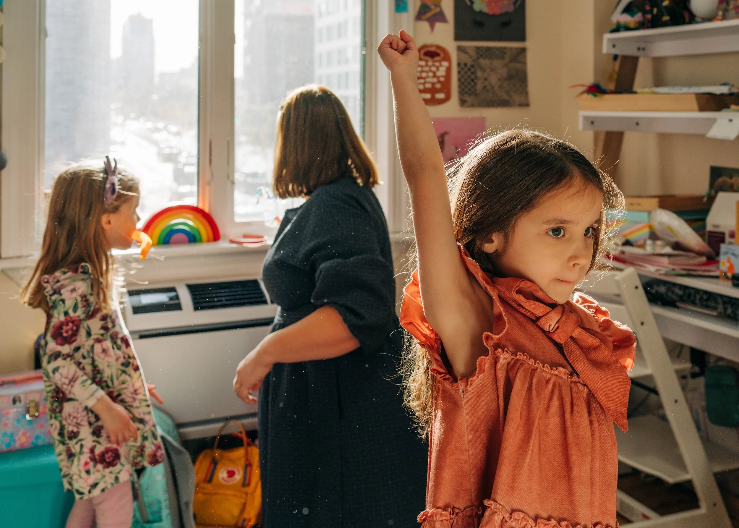 A young girl with long brown hair and a orange dress raising her arm inside a room with two other girls, books, and toys, and a window showing a cityscape in the background.