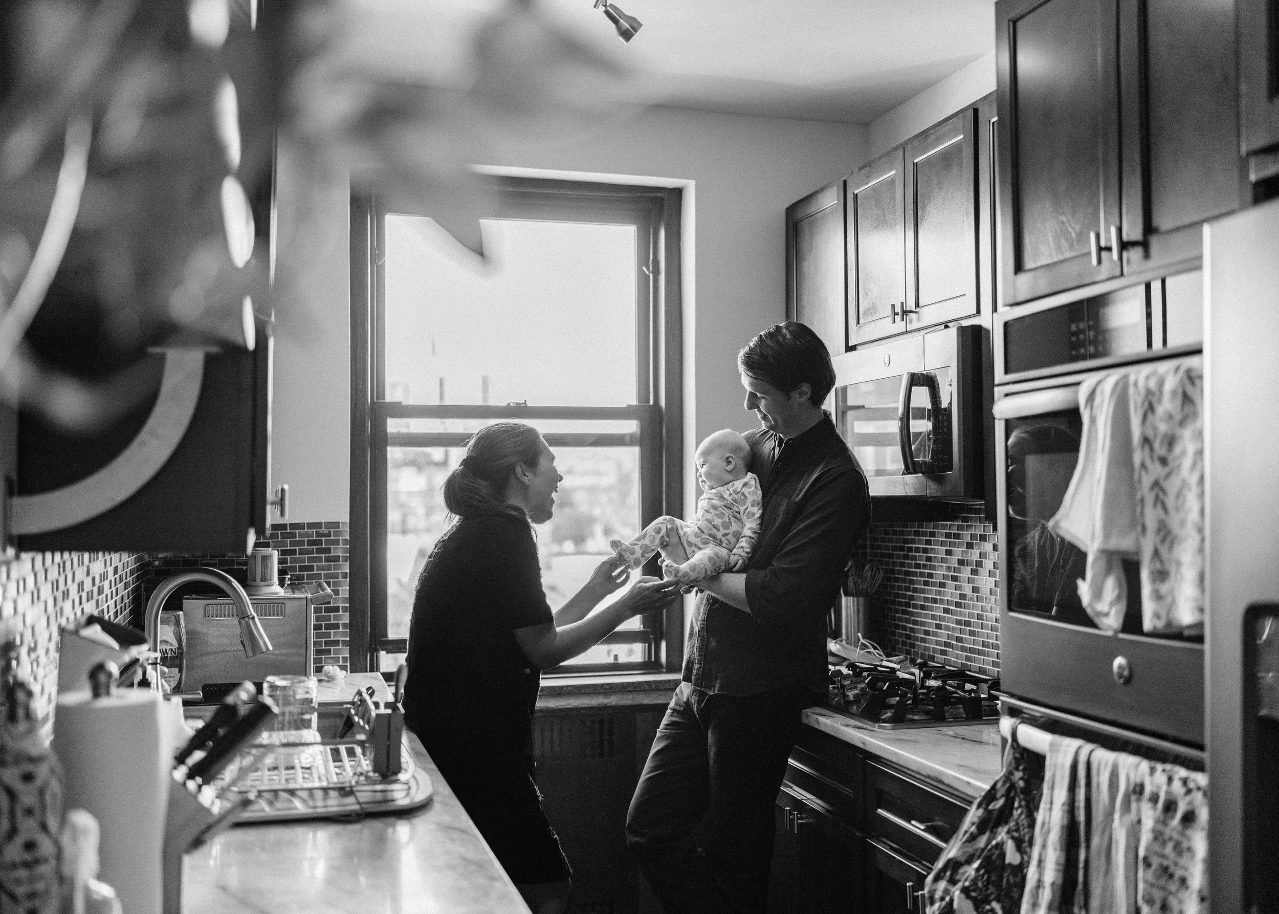 A woman and a man, possibly new parents, are in a kitchen. The woman is smiling and holding the baby's hand, while the man, holding a baby, looks at her. The kitchen has wooden cabinets, a window, and various appliances.