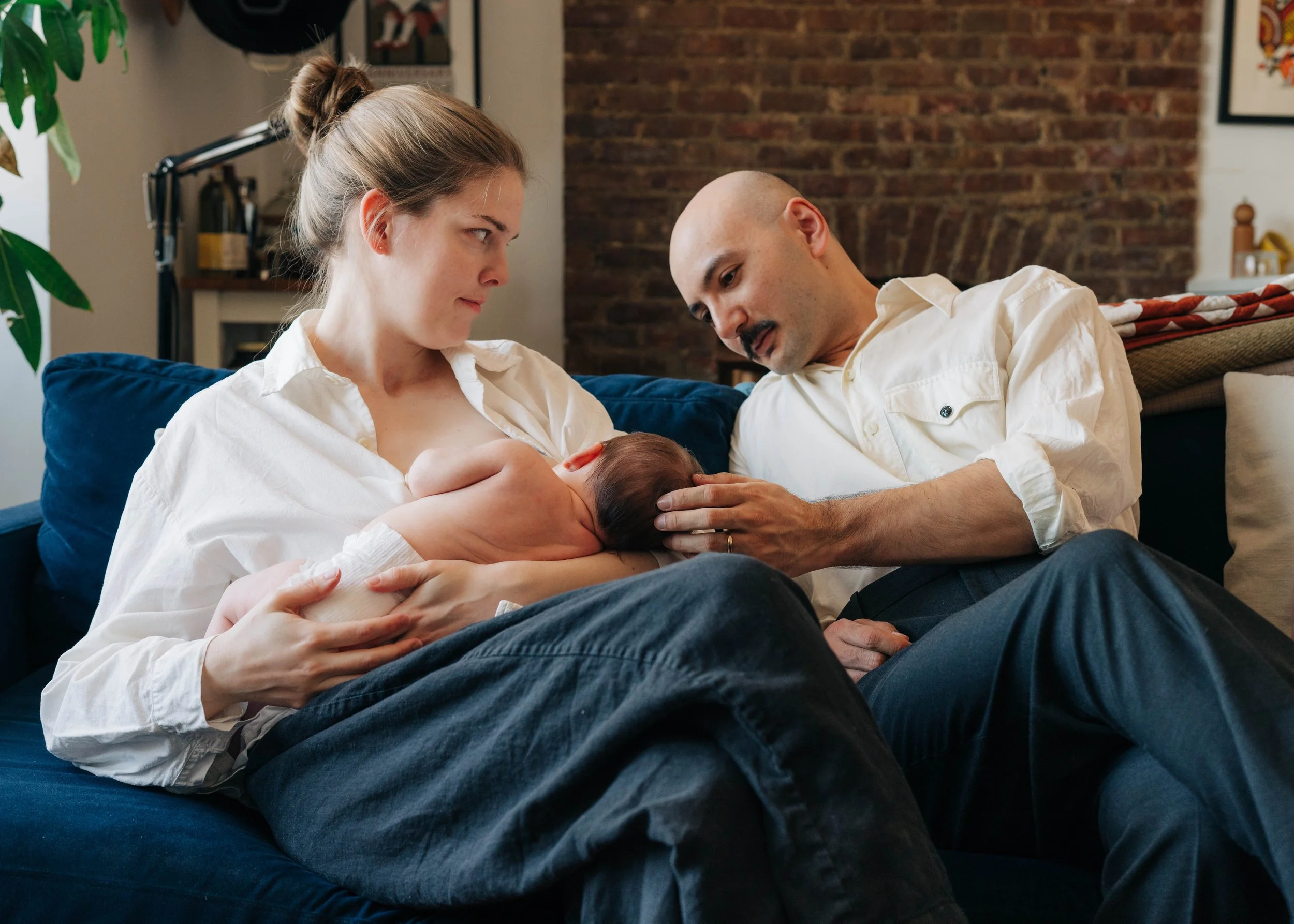 A woman and a man sit on a couch, looking at their newborn baby. The woman is holding the baby and looking at it intently, while the man gently touches the baby's head. The scene takes place in a cozy living room with a brick wall in the background.