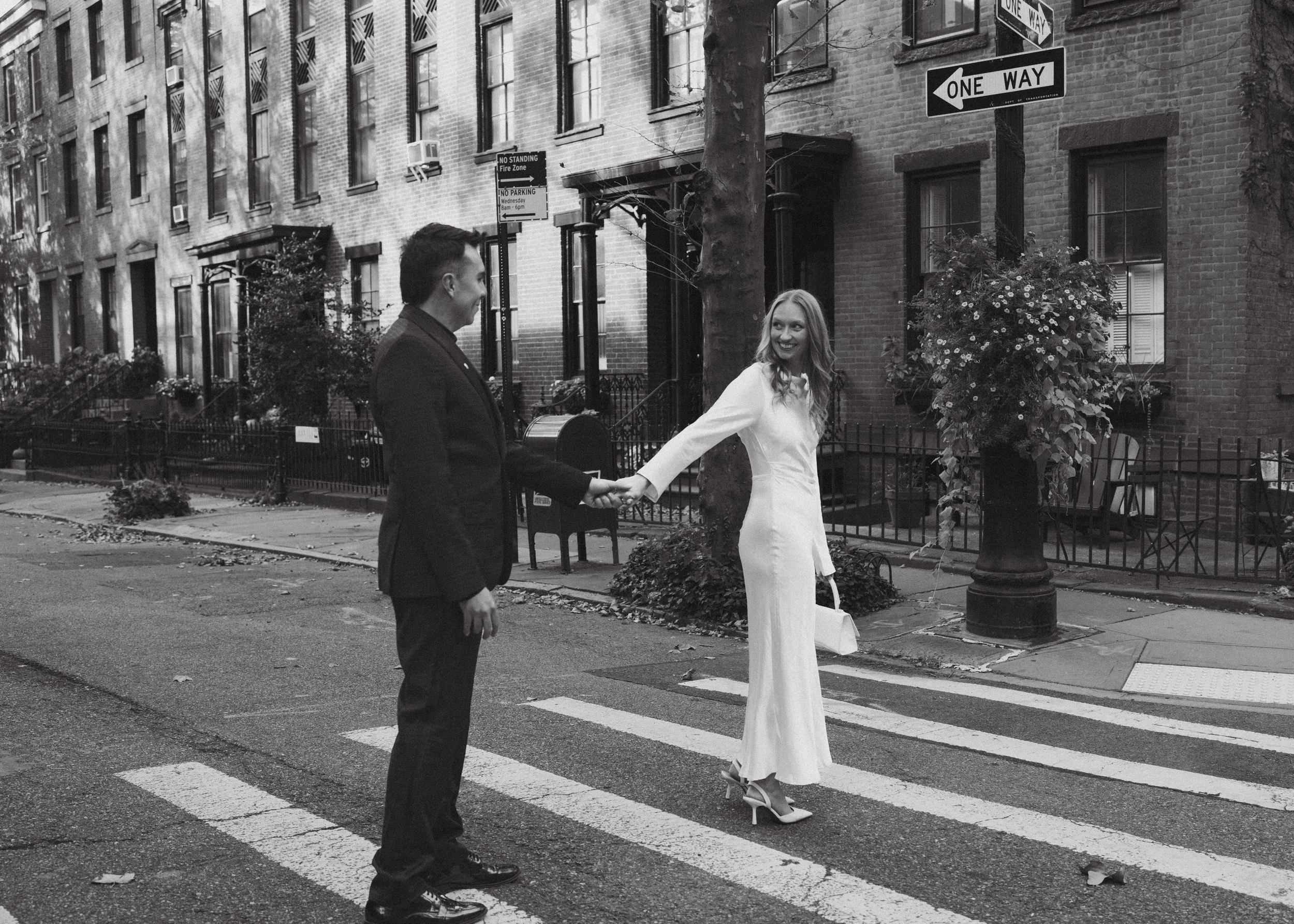 A black and white photo of a man and woman holding hands while crossing a street at a pedestrian crosswalk in an urban neighborhood. The man is dressed in a suit, and the woman is wearing a long dress and high heels, carrying a handbag. They are smil