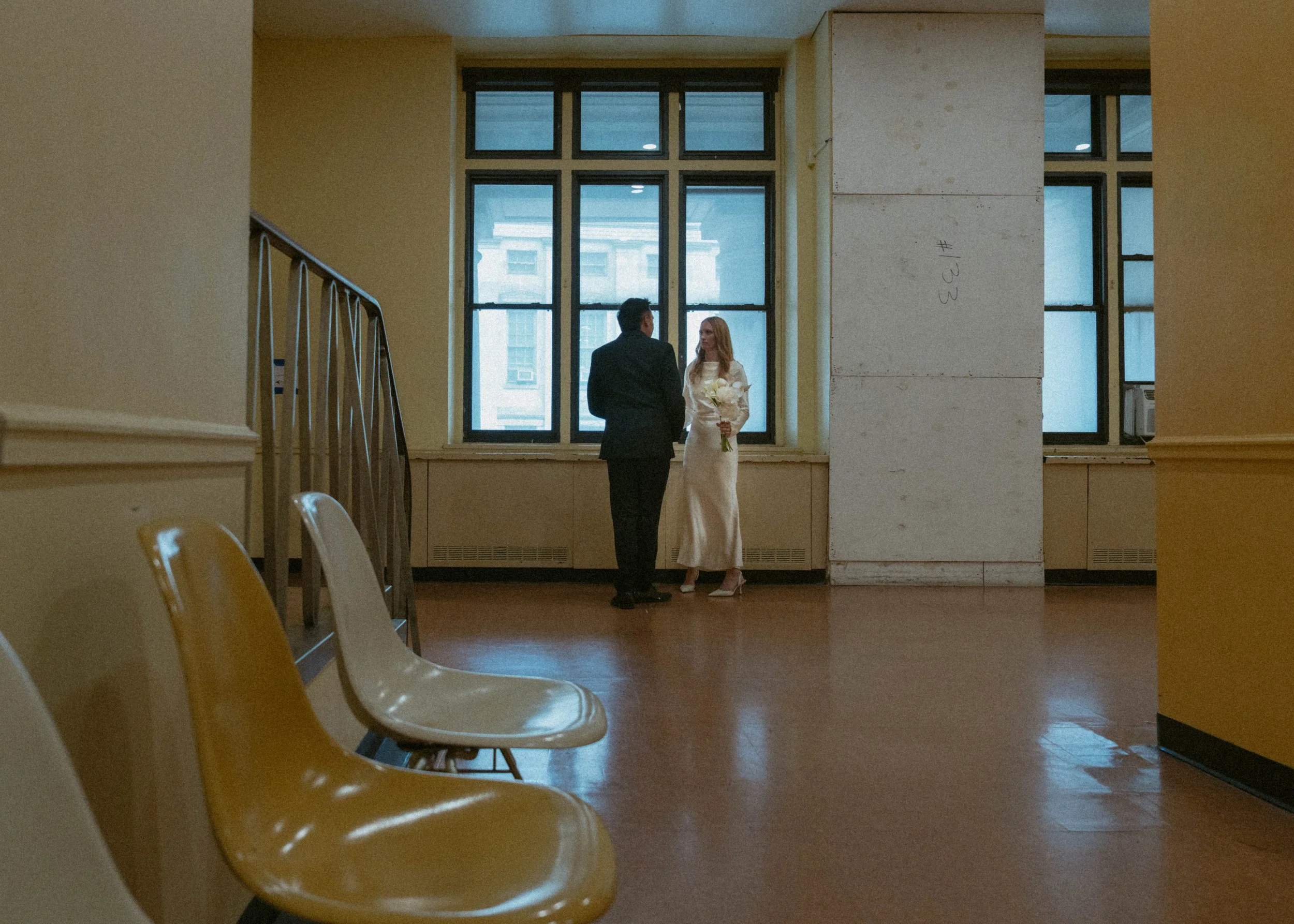 A bride in a white wedding dress holding a bouquet and a groom in a dark suit stand facing each other in front of large windows in a spacious room with yellow walls and empty yellow seats.