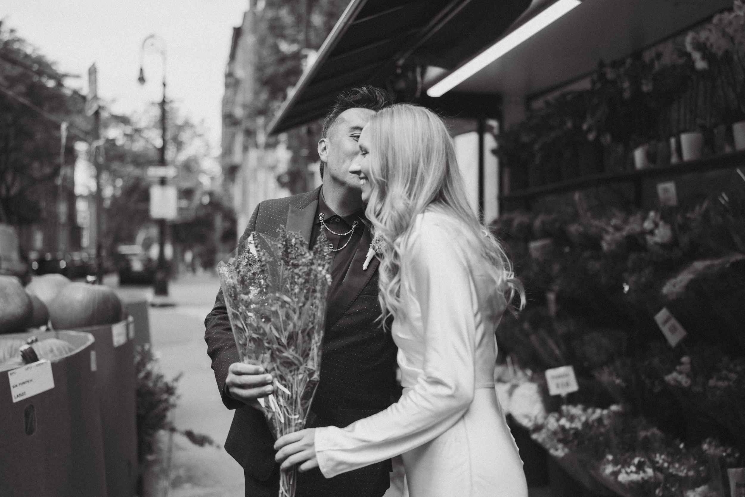 A man and woman sharing a kiss on a city sidewalk, the man holding a bouquet of flowers.