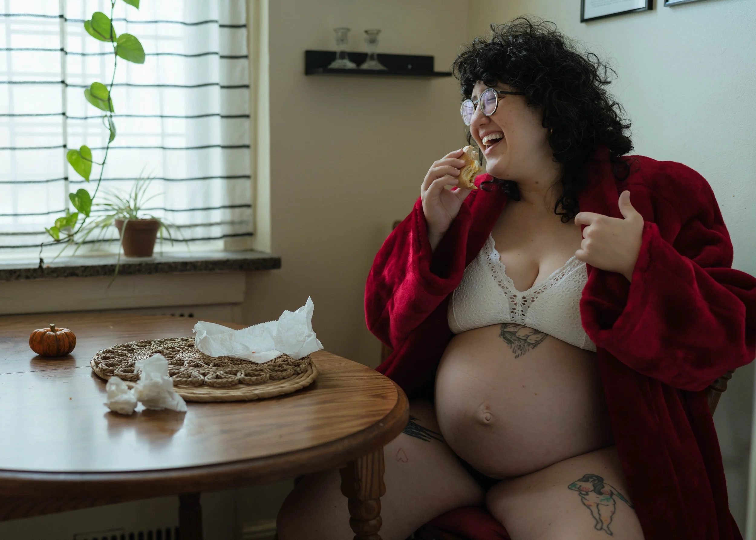 Pregnant woman with curly hair and glasses, sitting at a table, wearing a white lace bra and a red robe, smiling and eating a snack, with crumpled tissues on the table, small pumpkin, in a room with a window, plants, and wall decor.