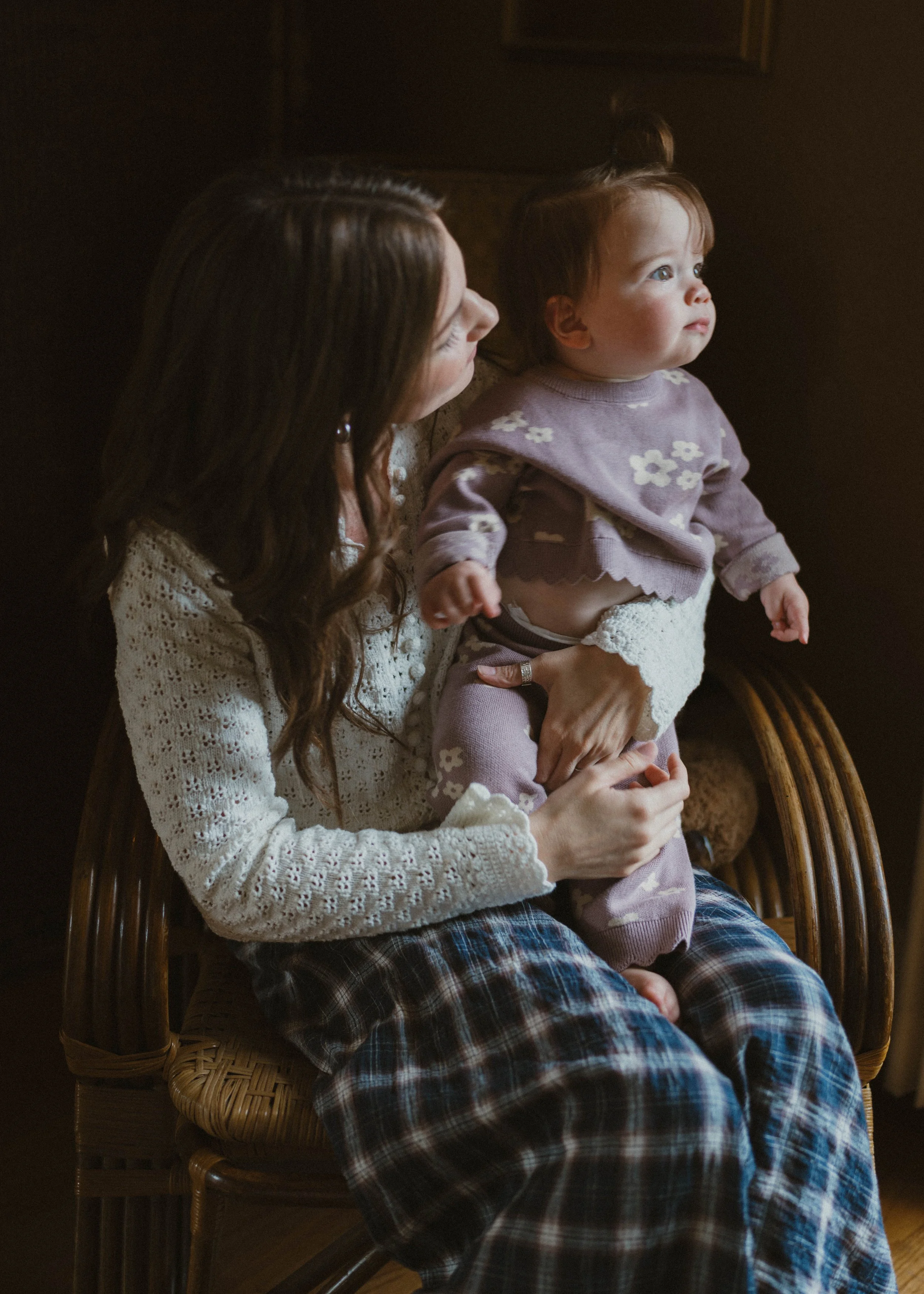 A woman with long brown hair holding a young girl with brown hair and eyes, sitting on a wooden chair indoors, lit by soft natural light.