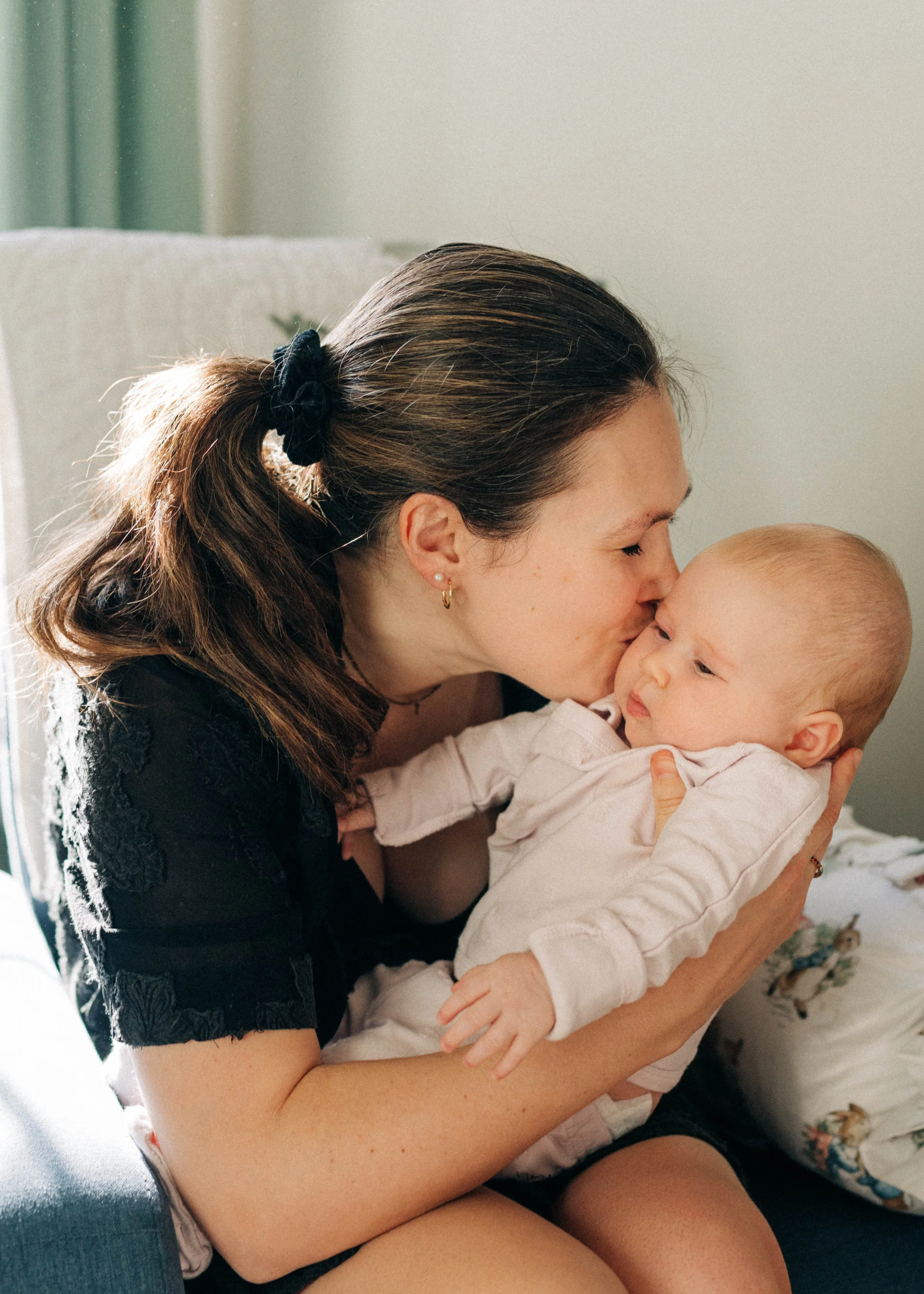 A woman with brown hair tied back with a black scrunchie, wearing earrings, kissing a baby on the forehead while holding the baby in her arms.