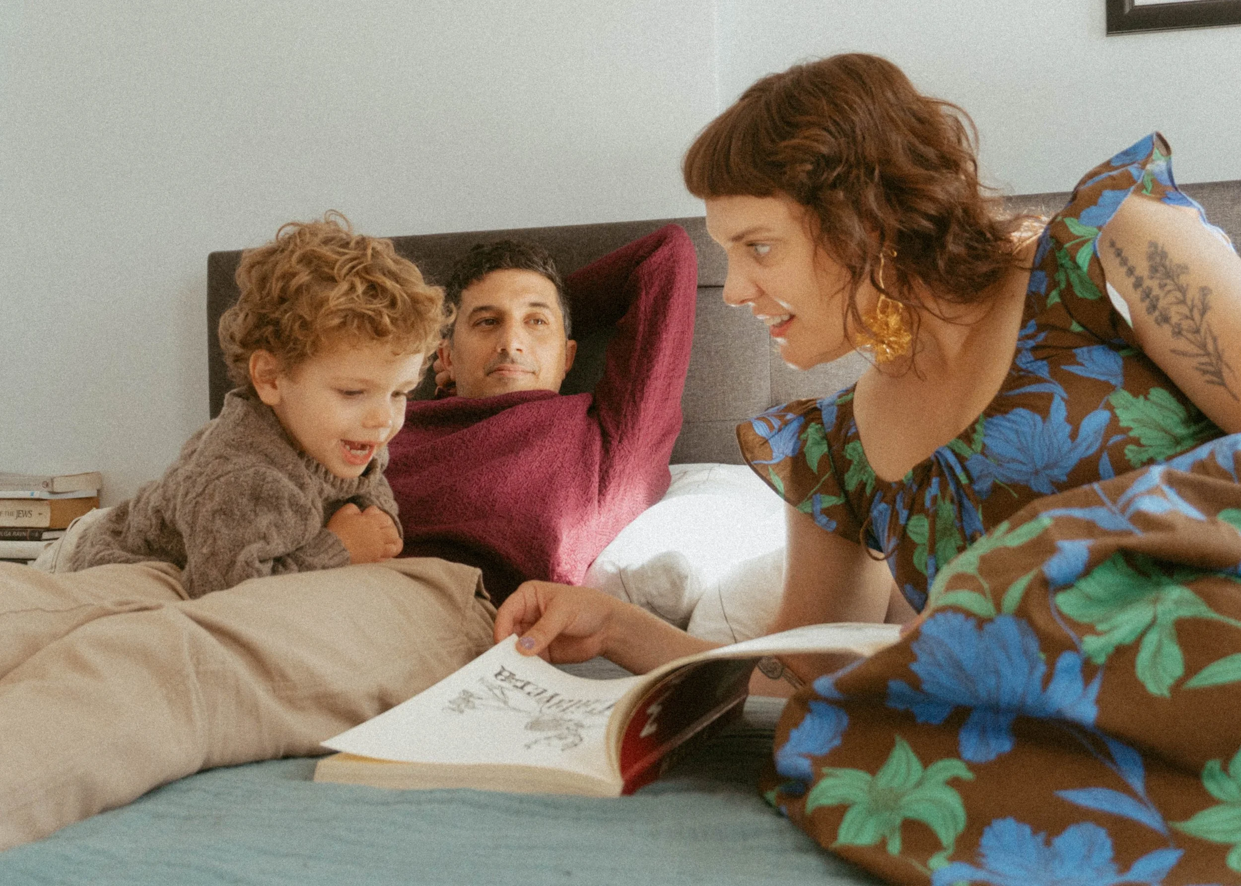 A woman is reading a book to a boy and a man lying on a bed. The woman has curly red hair and is wearing a floral dress, while the boy has curly hair and is wearing a cozy brown sweater. The man is lying back with his hands behind his head, smiling a