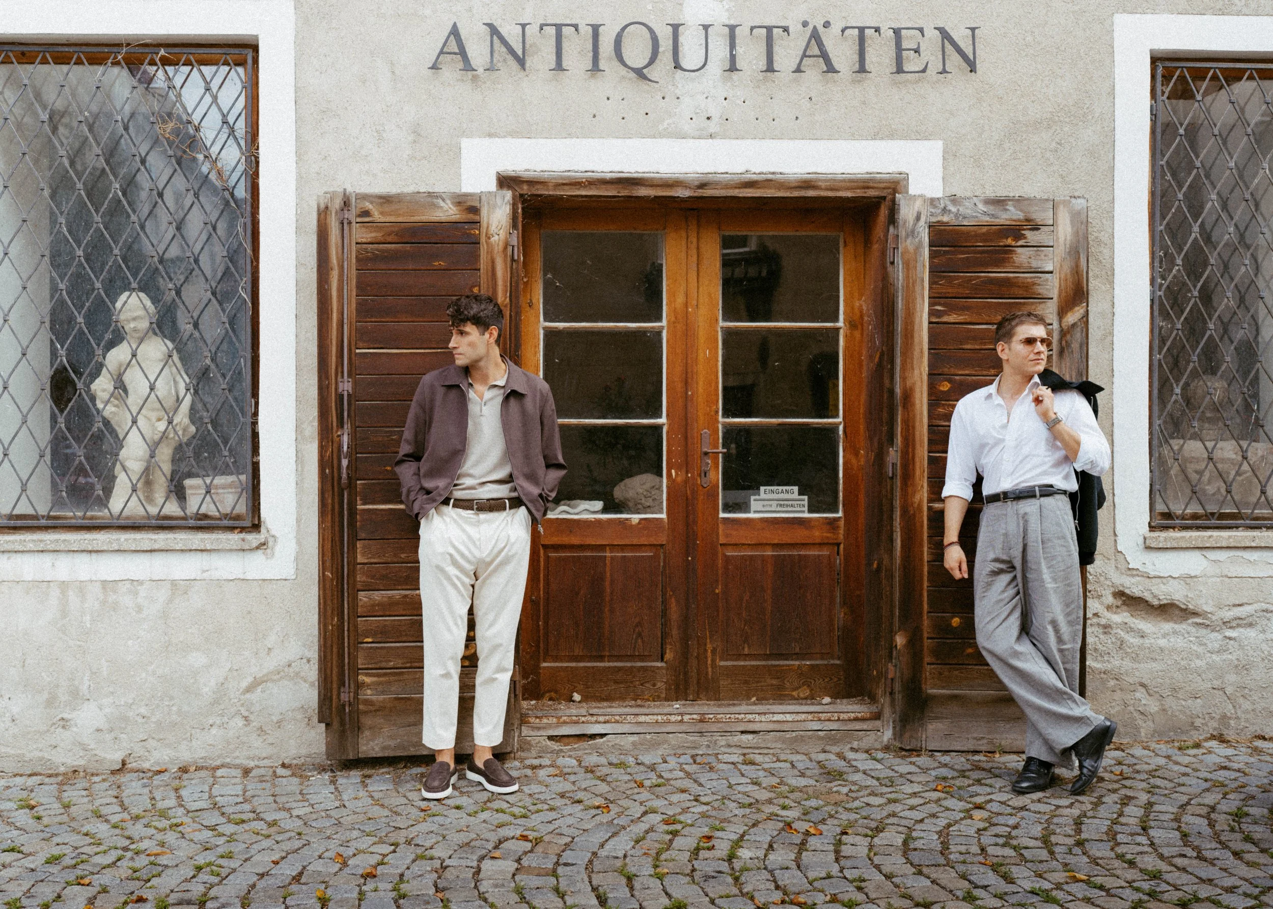 Two men stand outside a building with a sign reading 'Antiquitäten'. One man leans against the doorframe with his hands in his pockets, wearing a brown jacket and light-colored pants. The other man stands to the right with sunglasses, holding a jacket over his shoulder and dressed in a white shirt and gray pants.