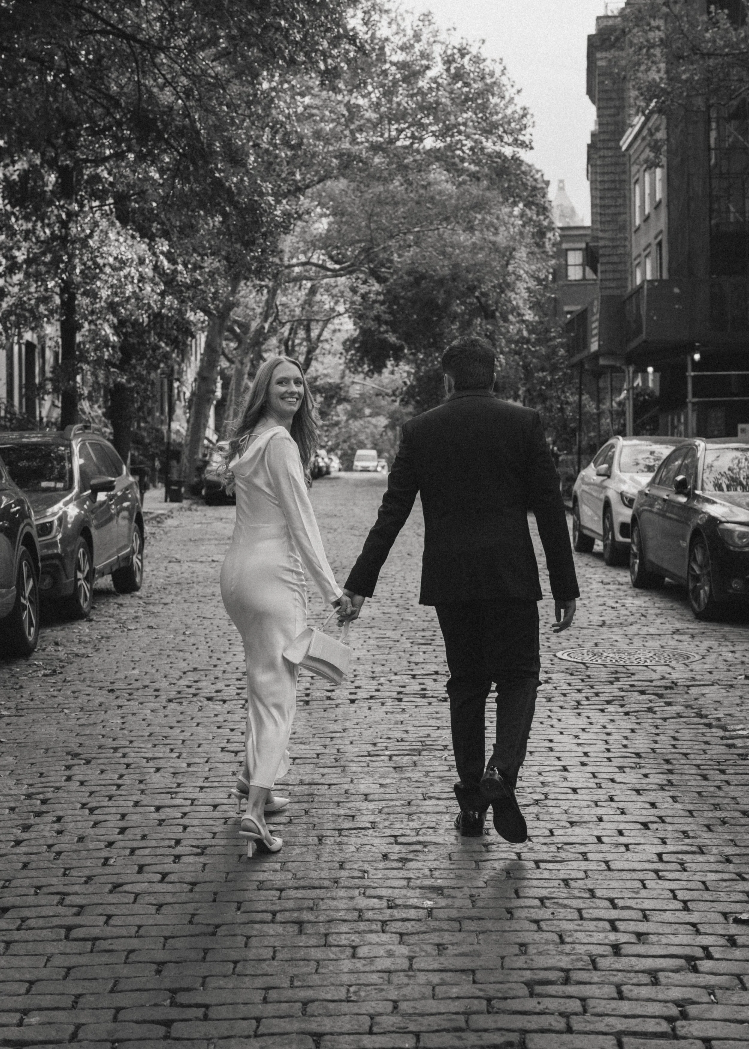 A black and white photo of a smiling woman in a satin dress and heels, holding a handbag, walking hand-in-hand with a man in a suit on a cobblestone street lined with parked cars and trees.