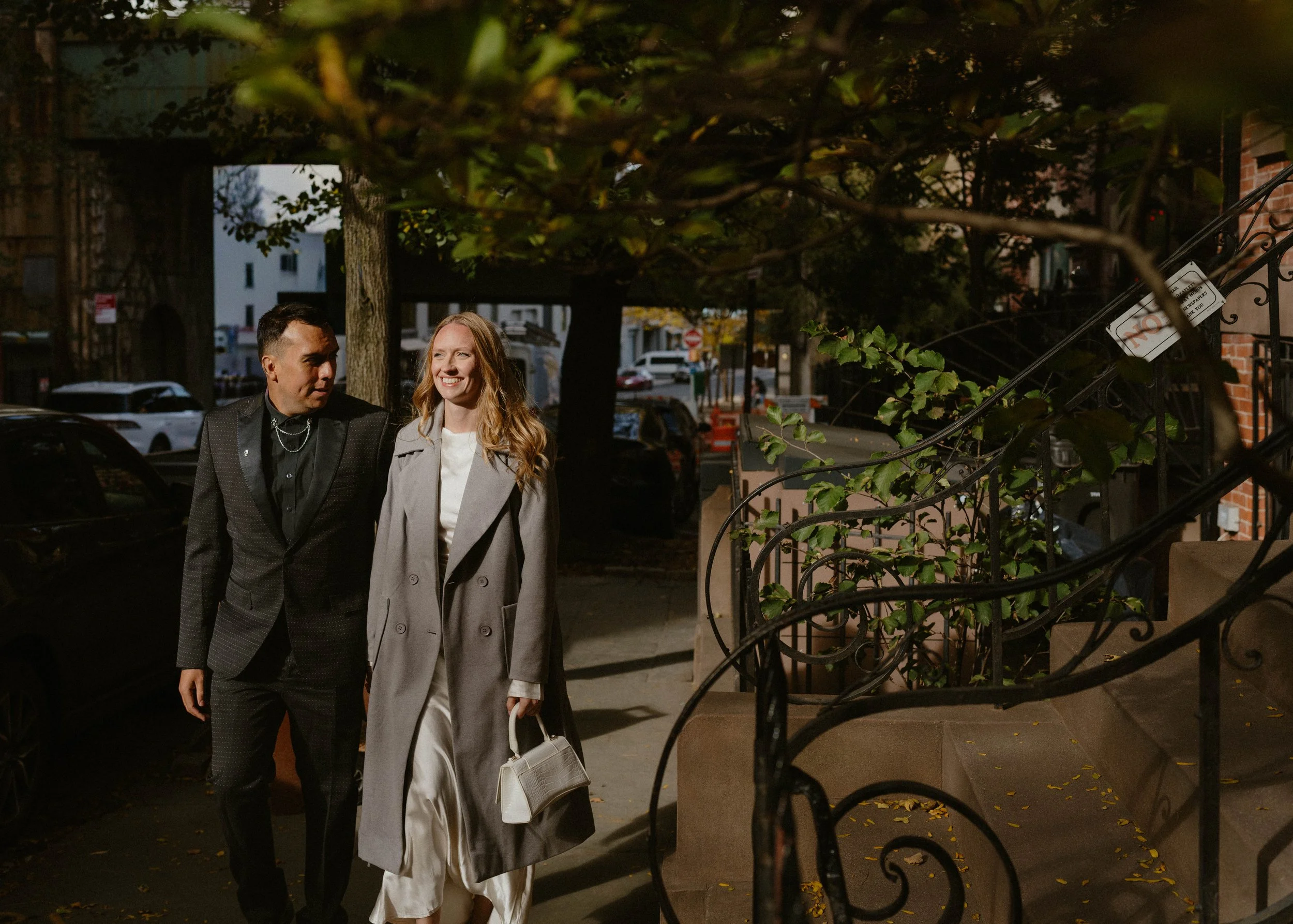 A man and a woman walking on a city sidewalk at dusk, smiling and talking. The man is wearing a black suit with a shiny blazer and the woman is dressed in a long gray coat and white pants, holding a small white handbag.
